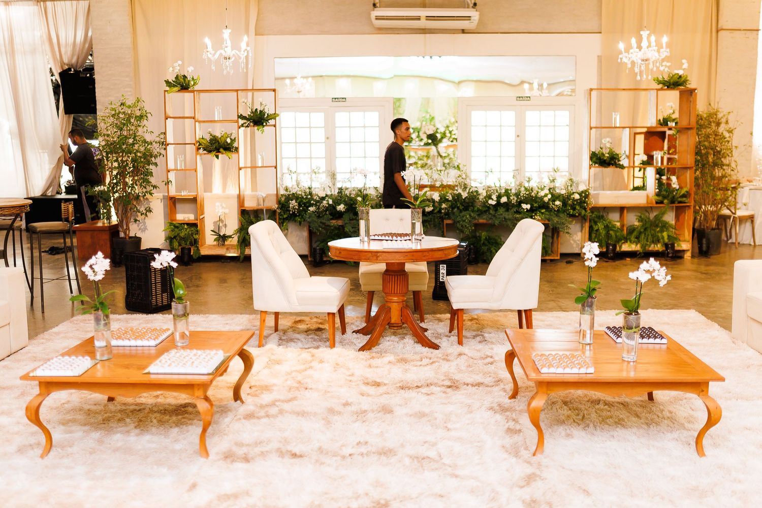 Elegant wedding reception lounge with white chairs, wooden tables, lush greenery, and a person standing near a floral backdrop.