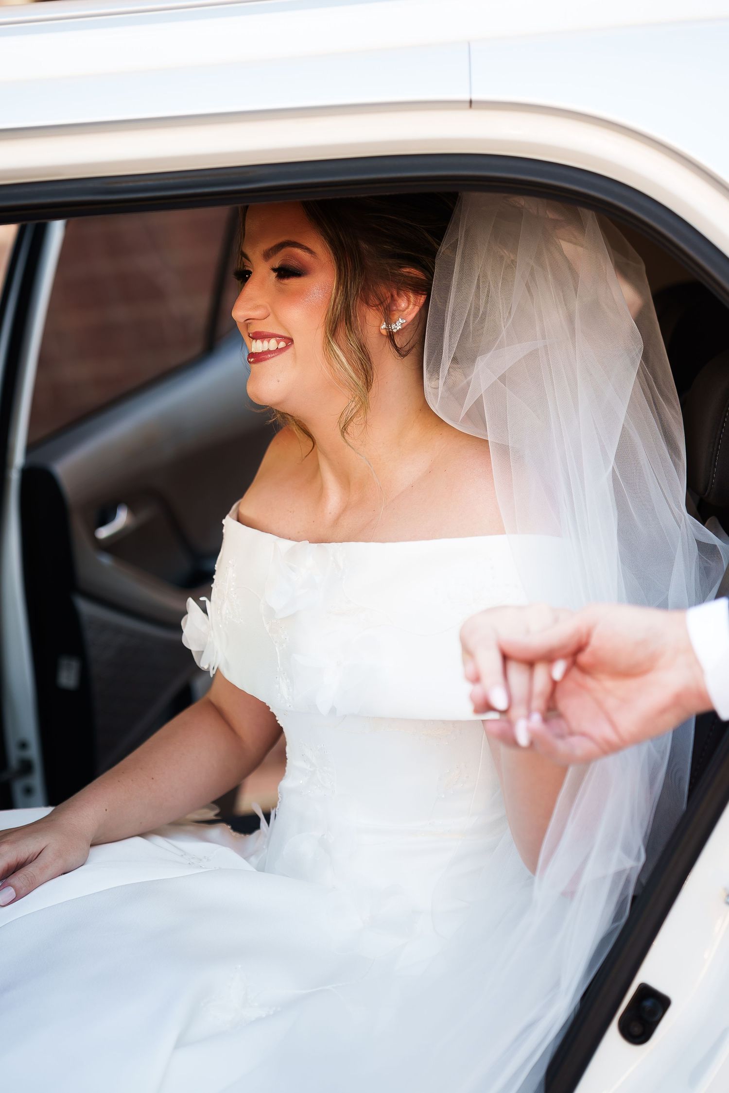 Smiling bride in white off-shoulder gown and veil sits in car, holding someone's hand on her wedding day.