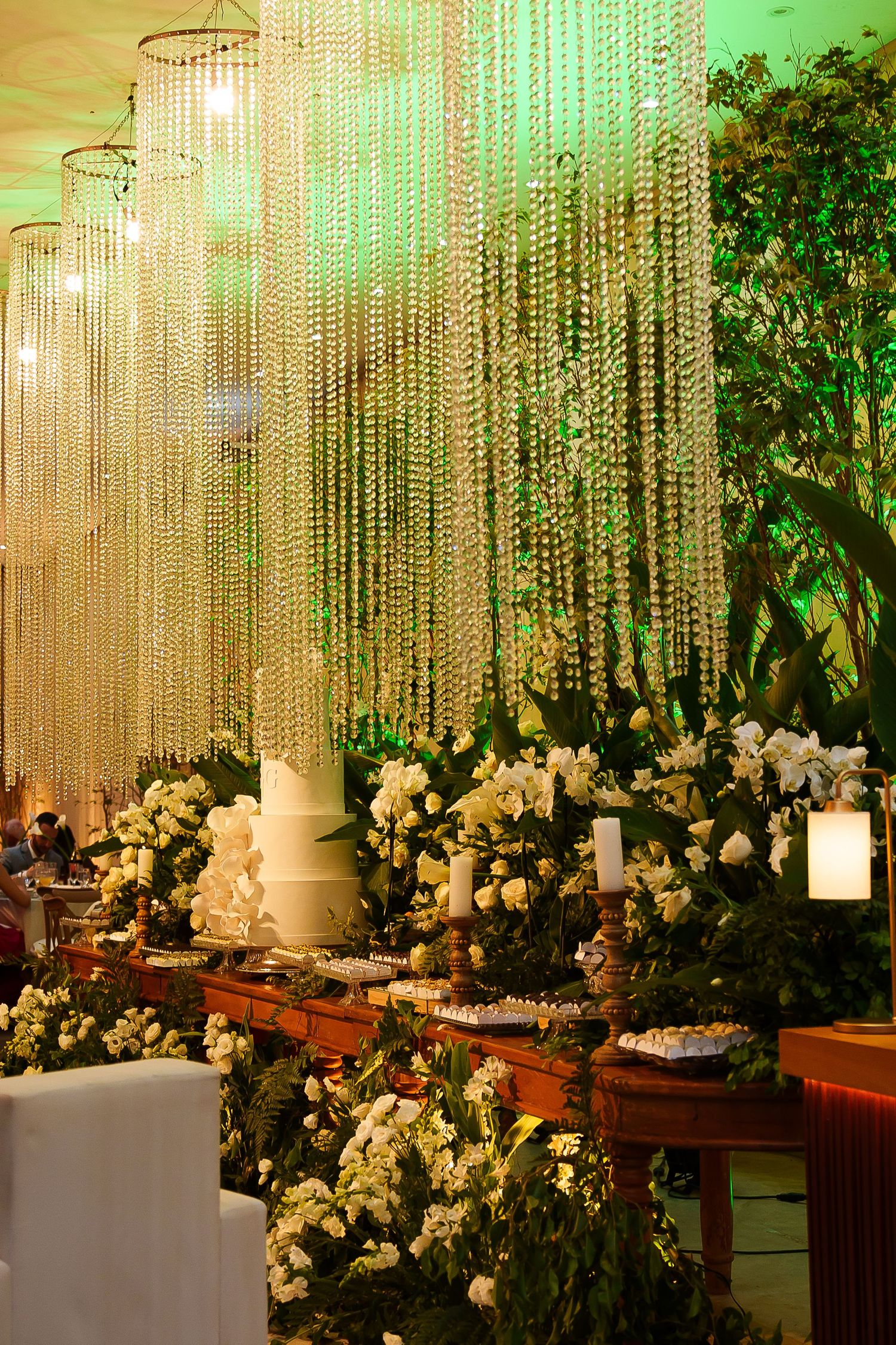 Elegant wedding reception with crystal bead chandeliers, white floral arrangements, and a tiered cake on a decorated table.