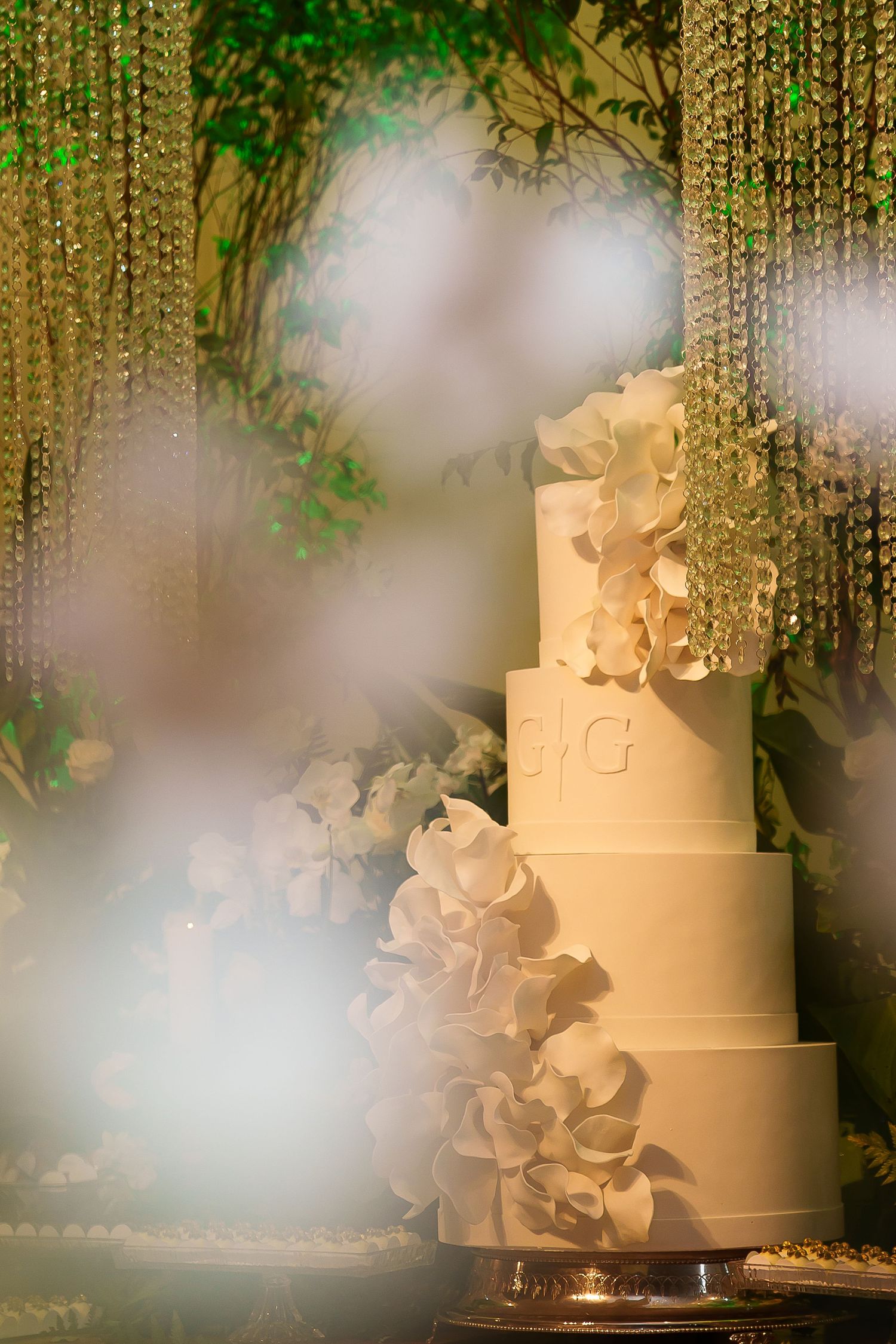 Elegant white multi-tier wedding cake adorned with orchids, monogram, crystal backdrop, and lush greenery.
