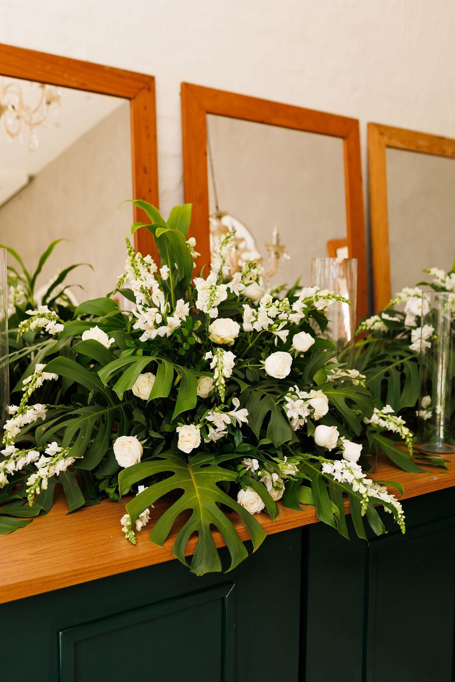 Elegant floral arrangement with white roses and monstera leaves on a wooden mantel with mirrors.
