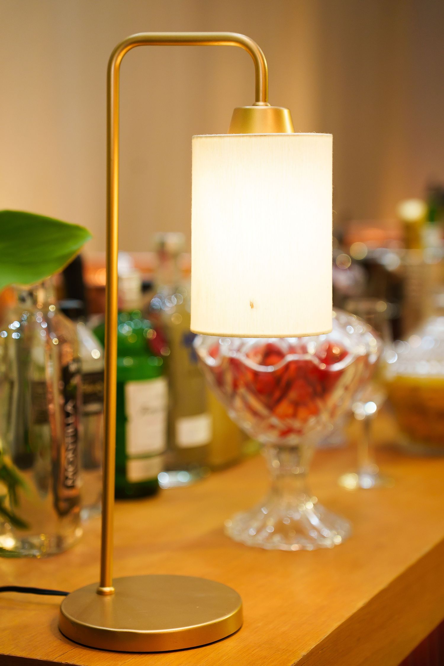 Gold arc desk lamp with white cylindrical shade on wooden bar counter with crystal glassware and bottles.
