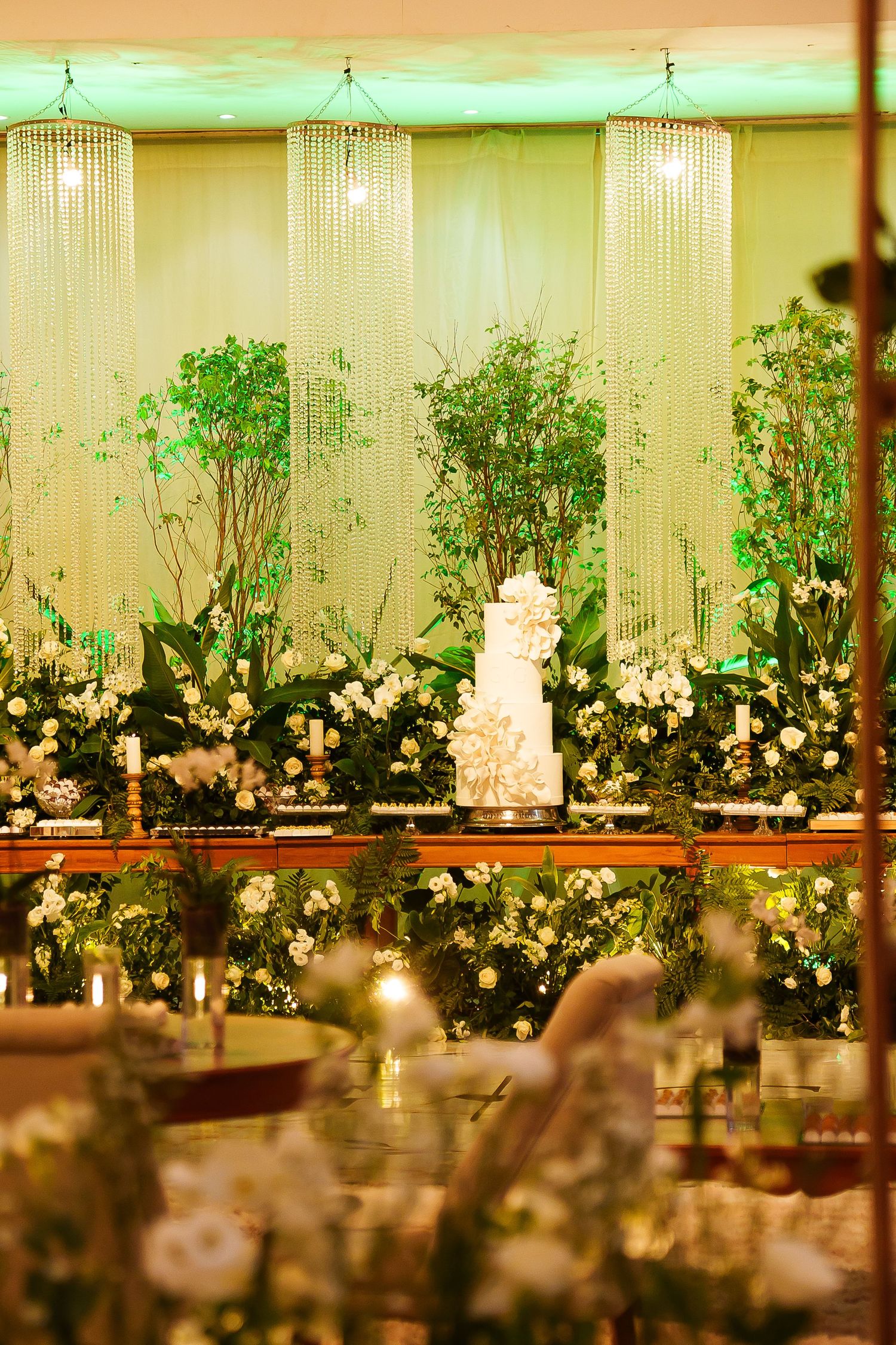 Elegant wedding reception with a white tiered cake surrounded by lush greenery, white flowers, and crystal chandeliers.