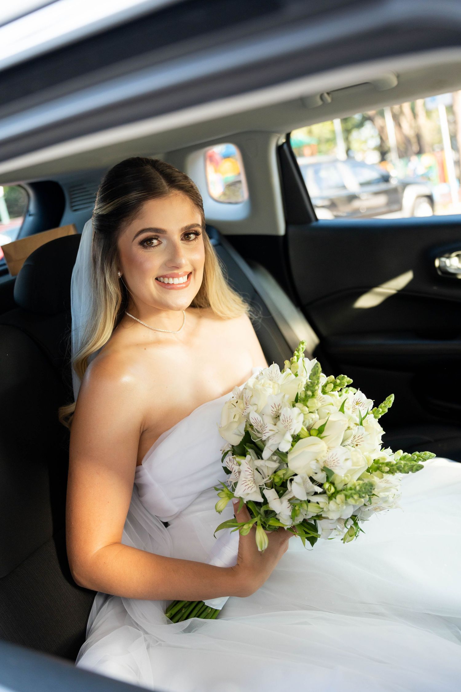 Smiling bride in white strapless gown holding bouquet of white flowers while sitting in car on wedding day.
