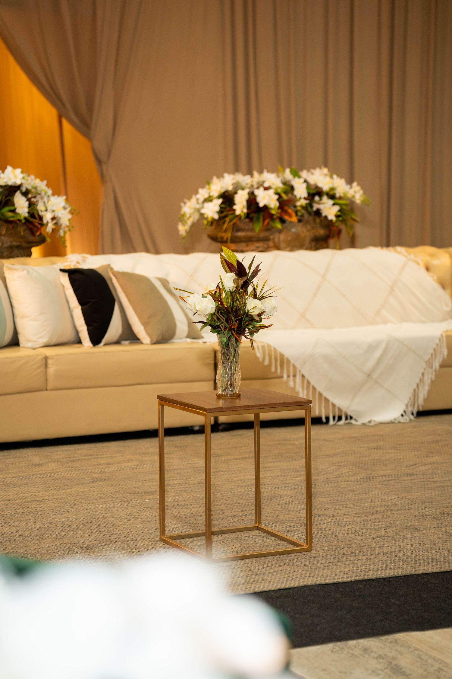 Elegant event lounge with cream sofa, fringed throw, gold side table, and fresh floral arrangements against draped backdrop.