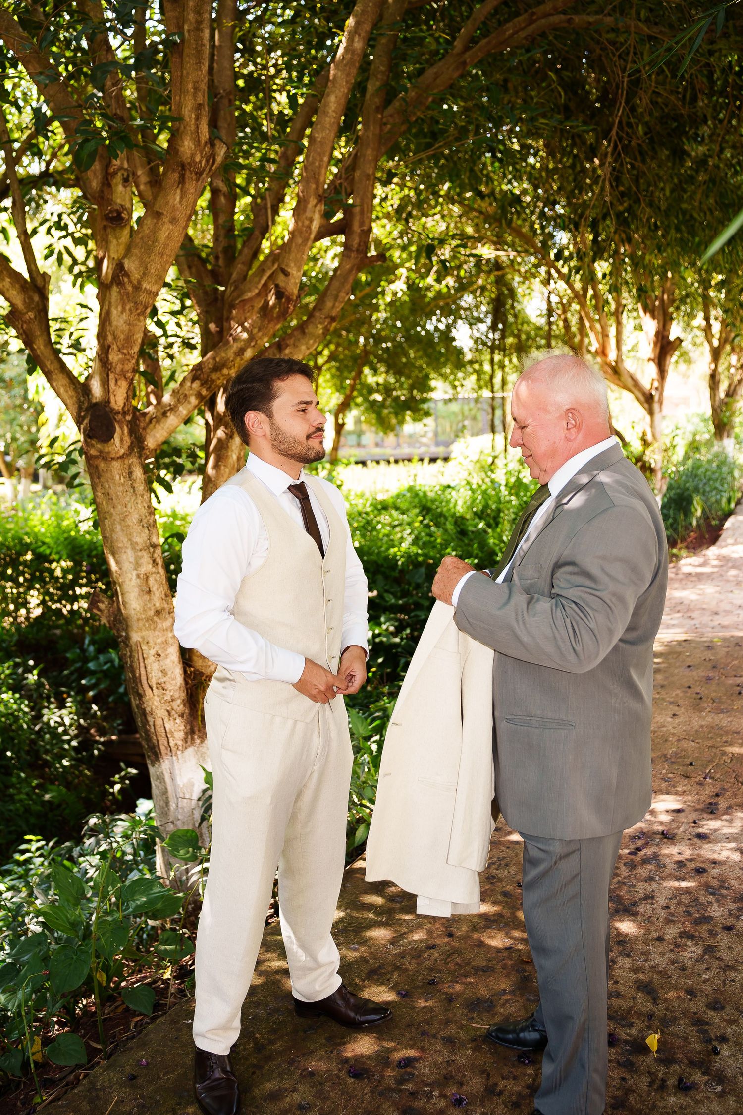 Groom in cream vest and pants receives his suit jacket from an older man in gray suit outdoors in a lush garden.