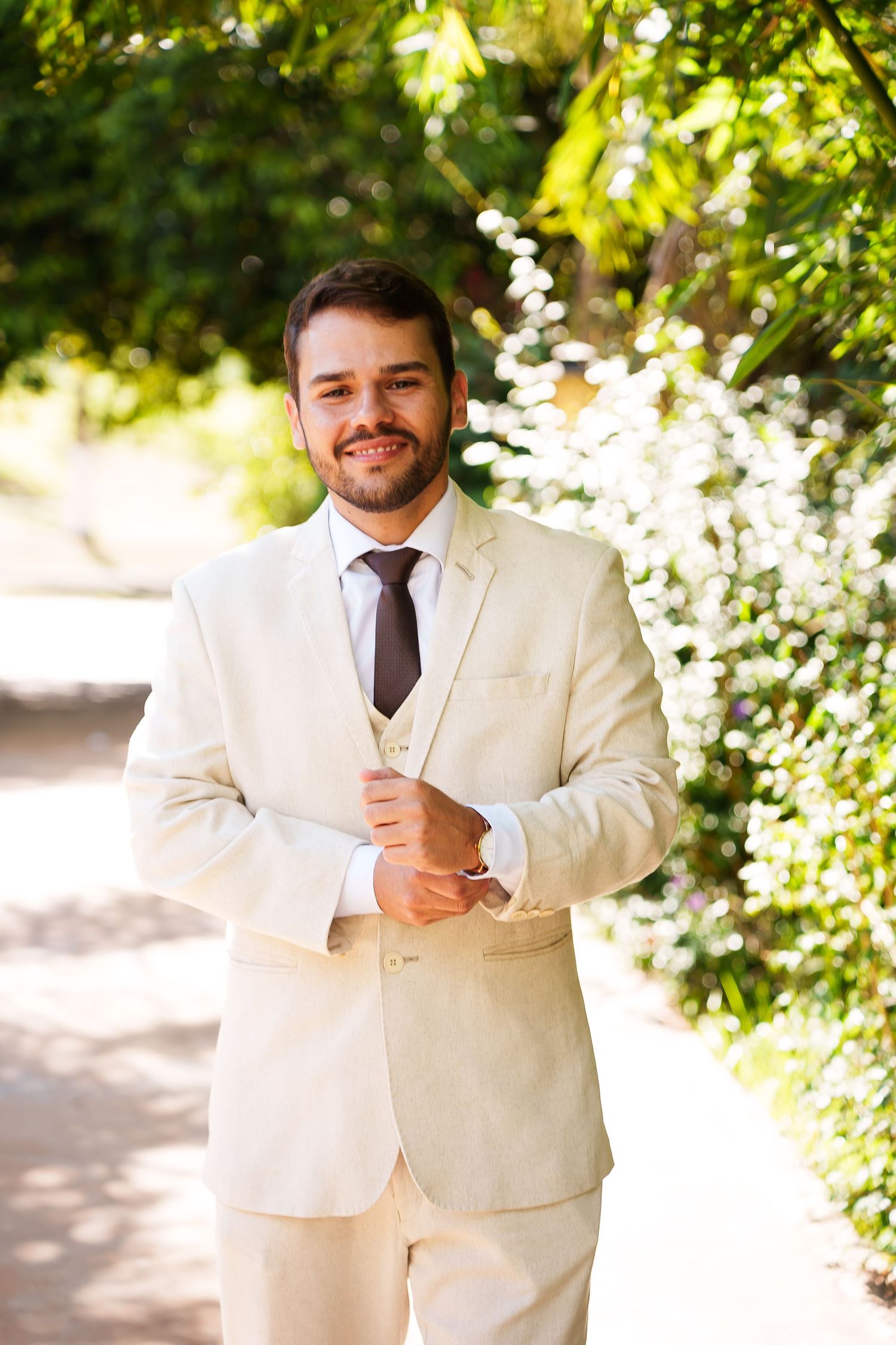 Smiling groom in cream suit adjusting cufflinks outdoors among lush green trees on wedding day.