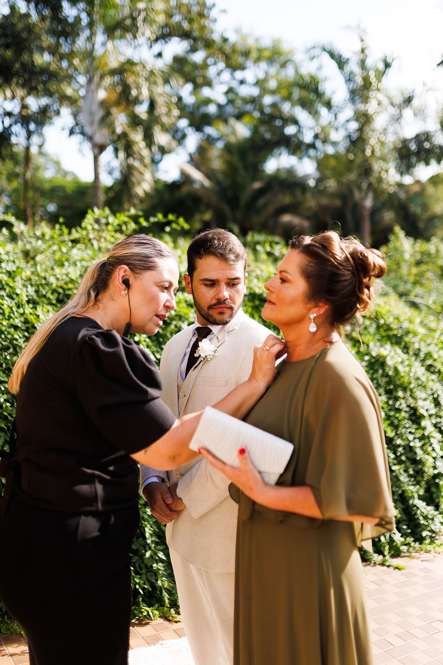 A wedding coordinator in black adjusts a grooms beige suit while speaking with an elegant woman in an olive dress outdoors.