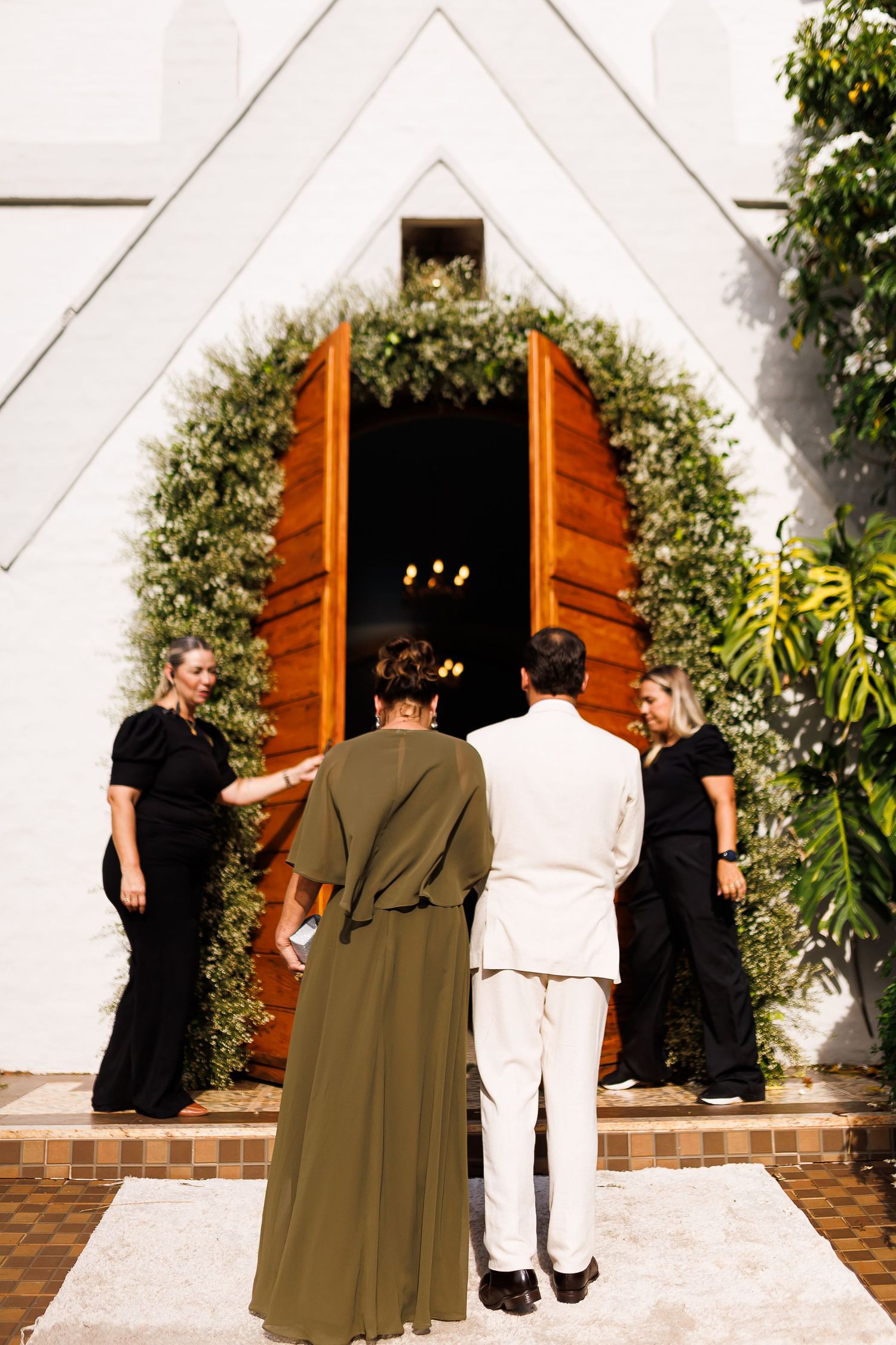 Couple entering a white chapel with wooden arched doors adorned with greenery, greeted by two women in black.