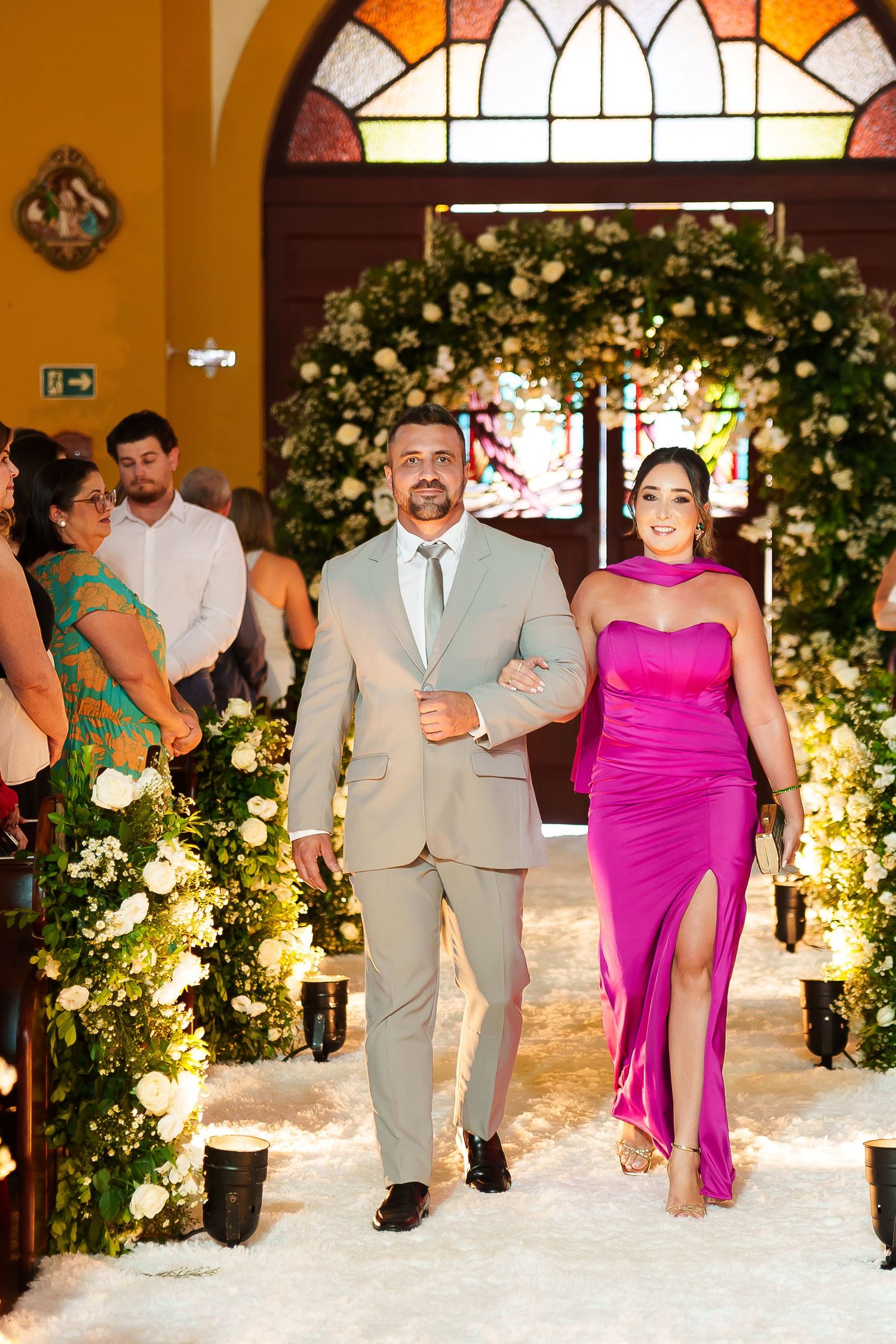 Couple walks down church aisle, man in beige suit, woman in pink gown, floral arch and candles in background.