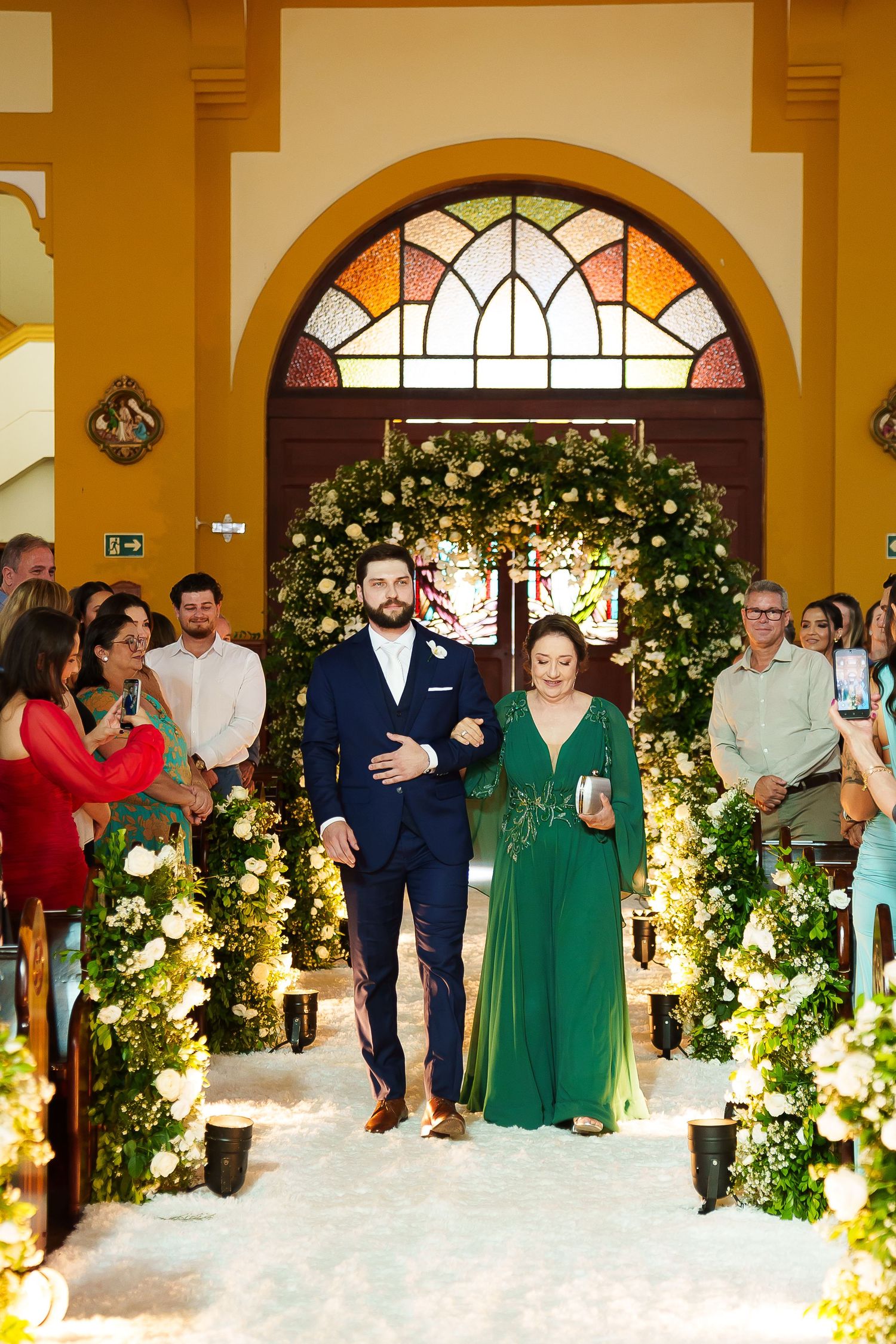 Groom in navy suit walks down church aisle with woman in green gown, flanked by floral arrangements and wedding guests.