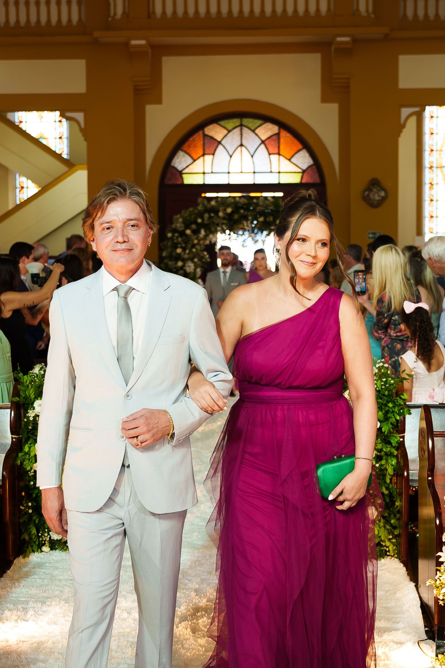 Elegant couple posing in church aisle, man in light gray suit, woman in magenta one-shoulder tulle gown holding green clutch.