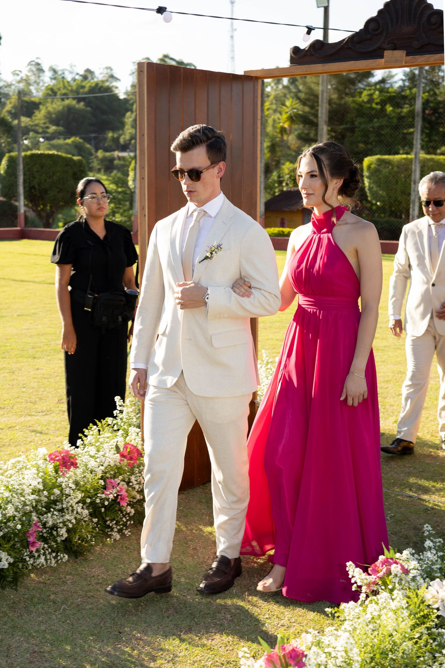 Couple in formal attire at outdoor wedding, man in cream suit, woman in red halter gown, floral decorations.
