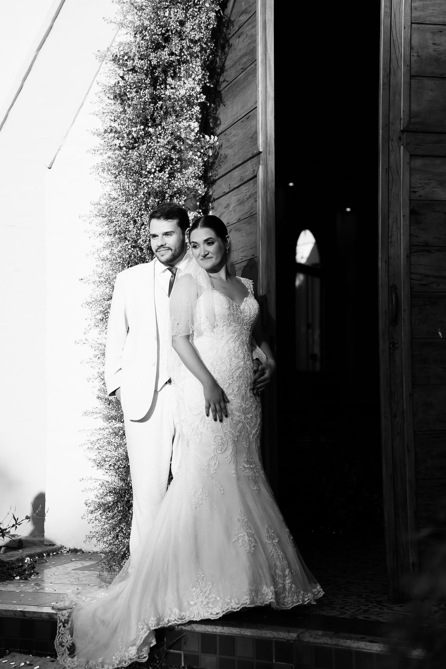 Black and white wedding photo of bride in lace gown and groom in white suit posing elegantly at a rustic doorway.