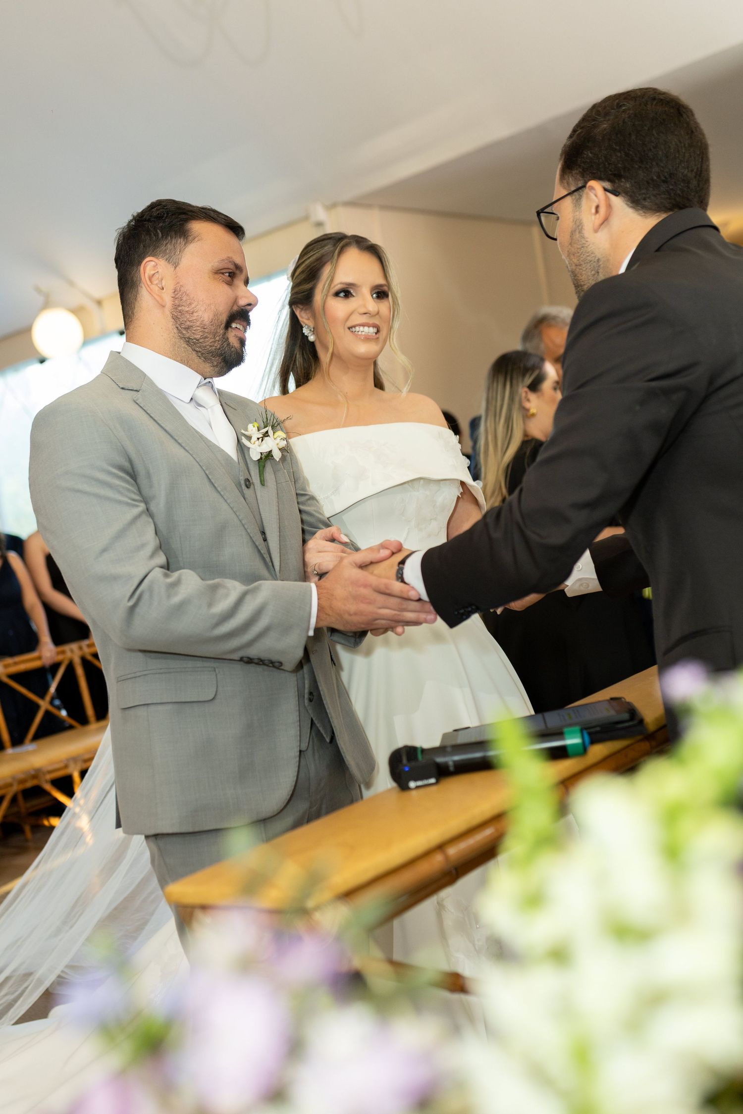 A bride and groom exchange vows at their wedding ceremony, smiling joyfully while an officiant holds their hands.