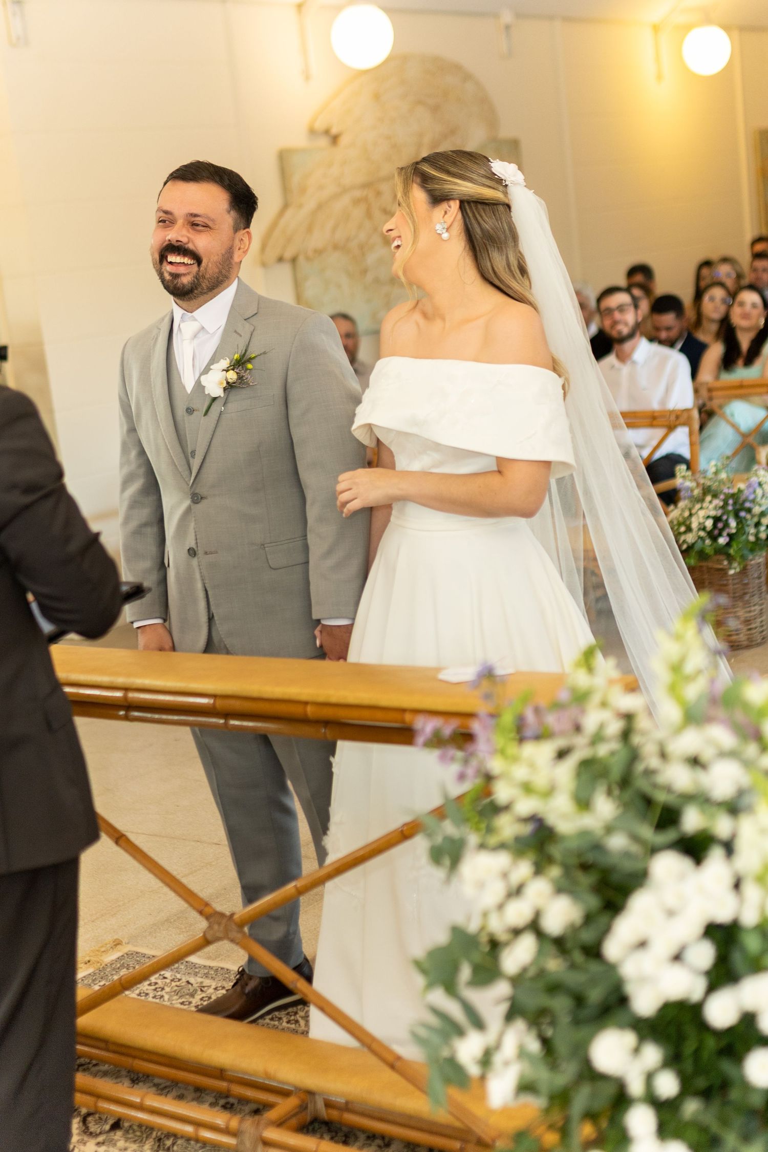 Bride in off-shoulder white gown and veil stands with smiling groom in gray suit at church altar ceremony.
