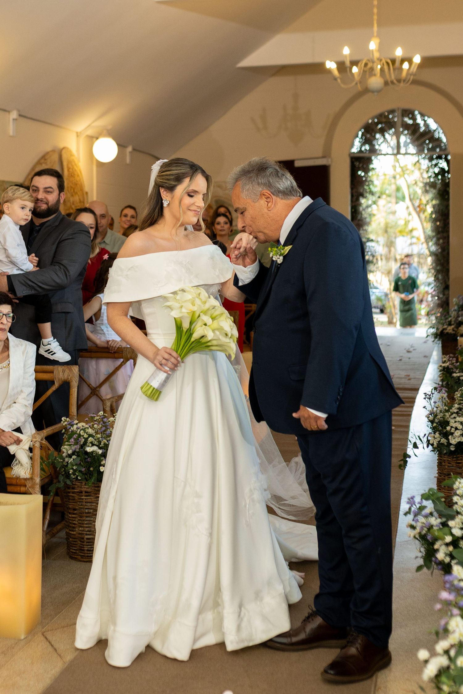 Bride in off-shoulder white gown holds calla lilies as older man in navy suit kisses her hand in church aisle.