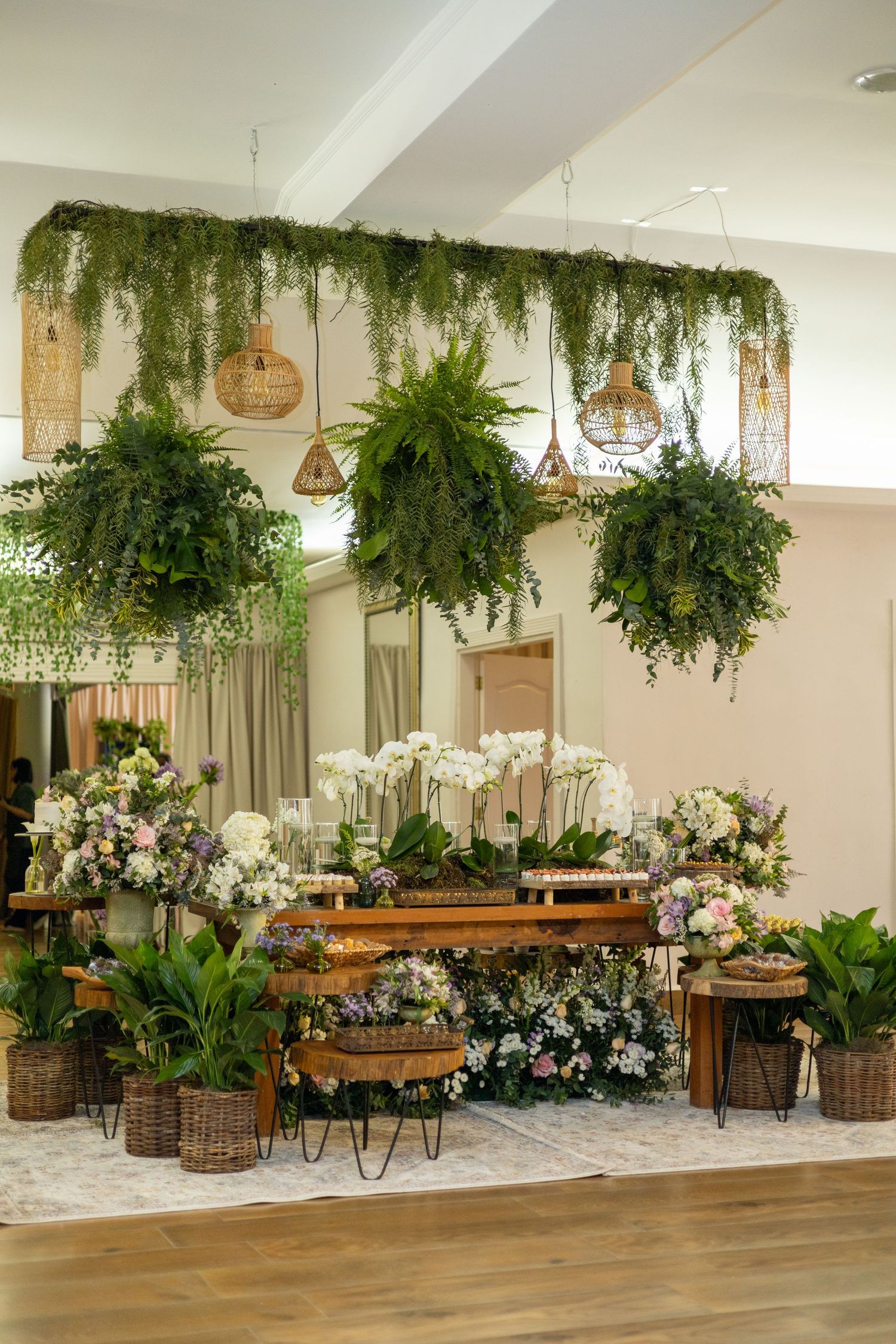 Lush indoor wedding display with hanging ferns, wicker pendants, and floral arrangements on rustic wooden tables.