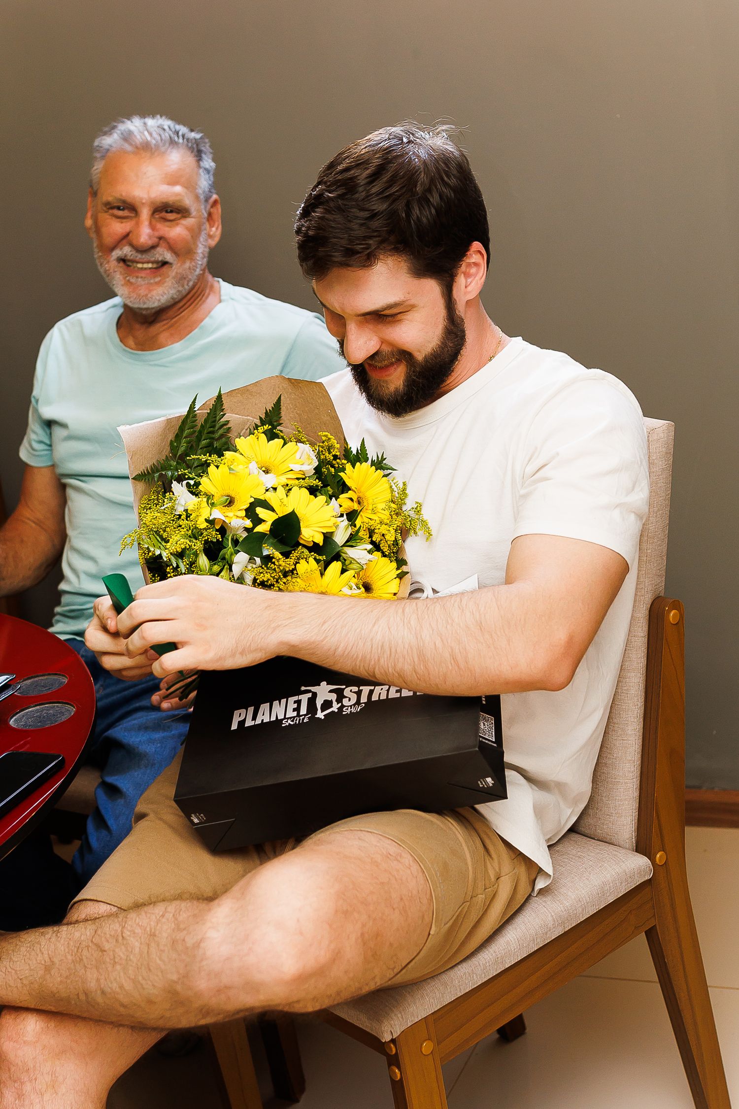 Young bearded man smiling while holding yellow flowers and a Planet Fitness box, with older man in background.