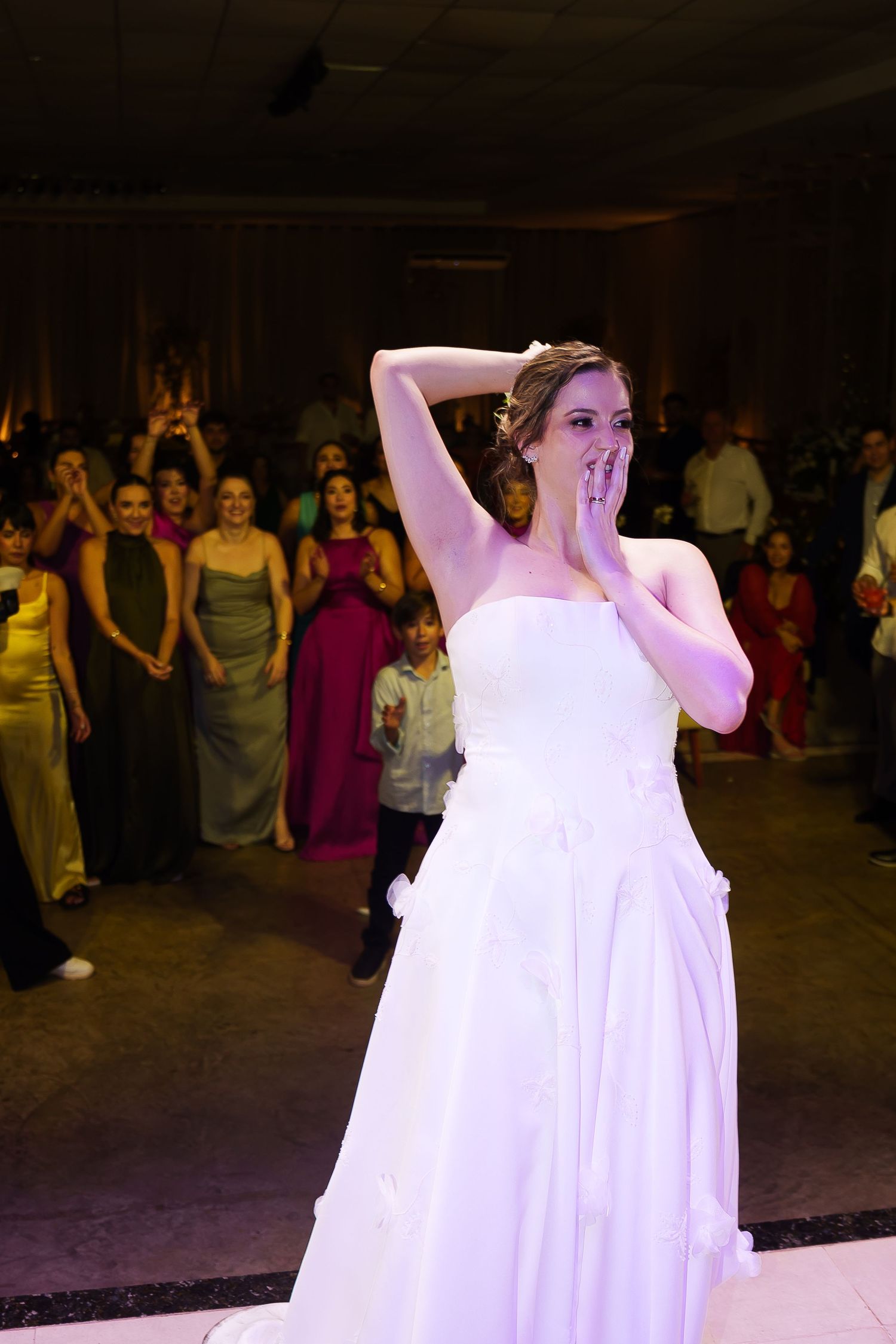 Bride in white strapless gown covers her mouth in surprise on wedding dance floor surrounded by guests.
