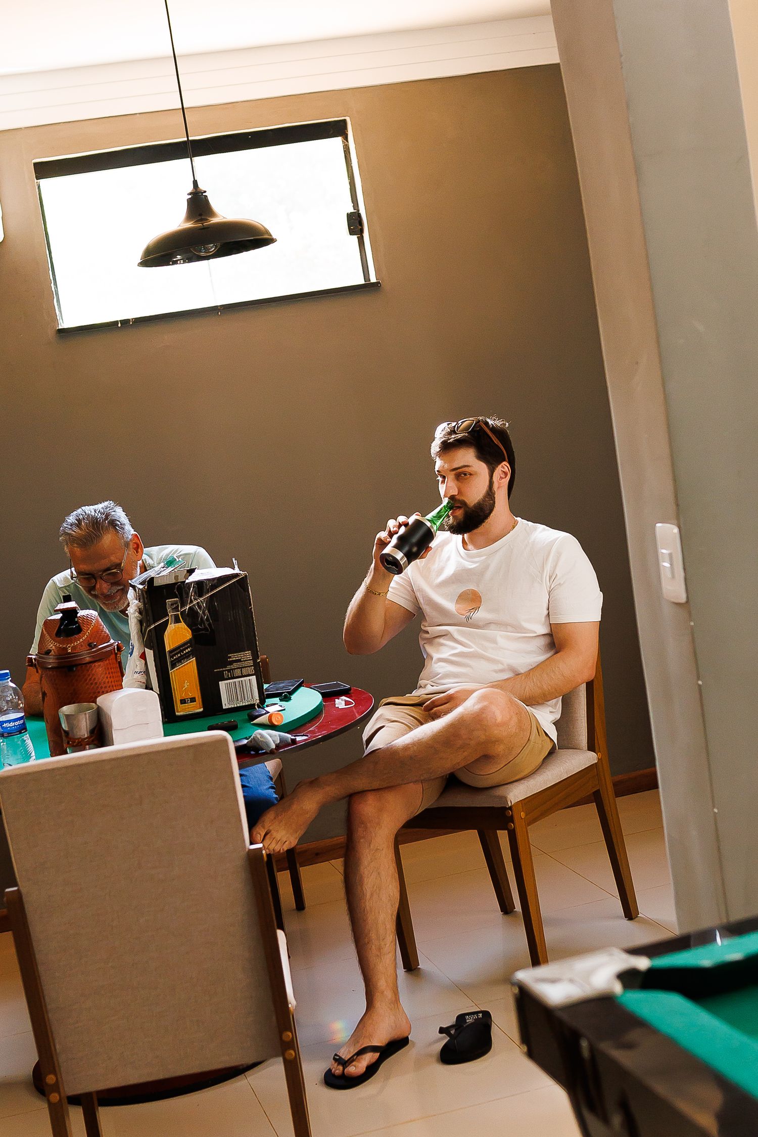 Two men relaxing indoors, one drinking a beer bottle while seated on a chair near a table with drinks.