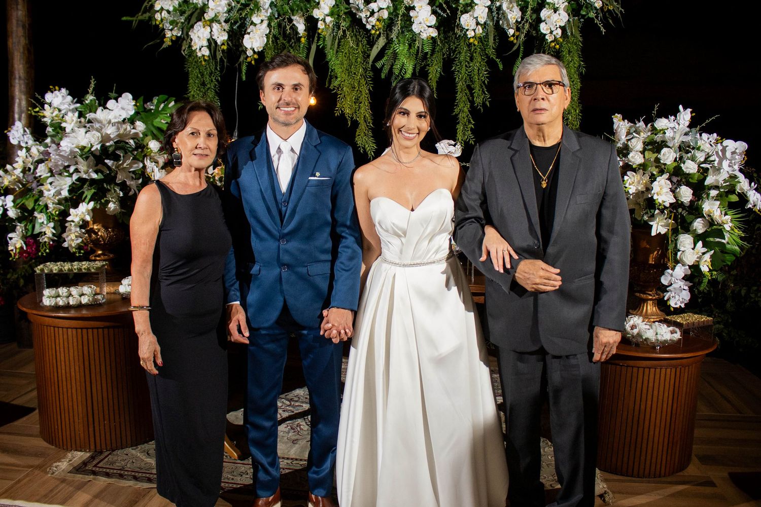 Wedding couple stands with family members in front of lush floral arrangement at reception.