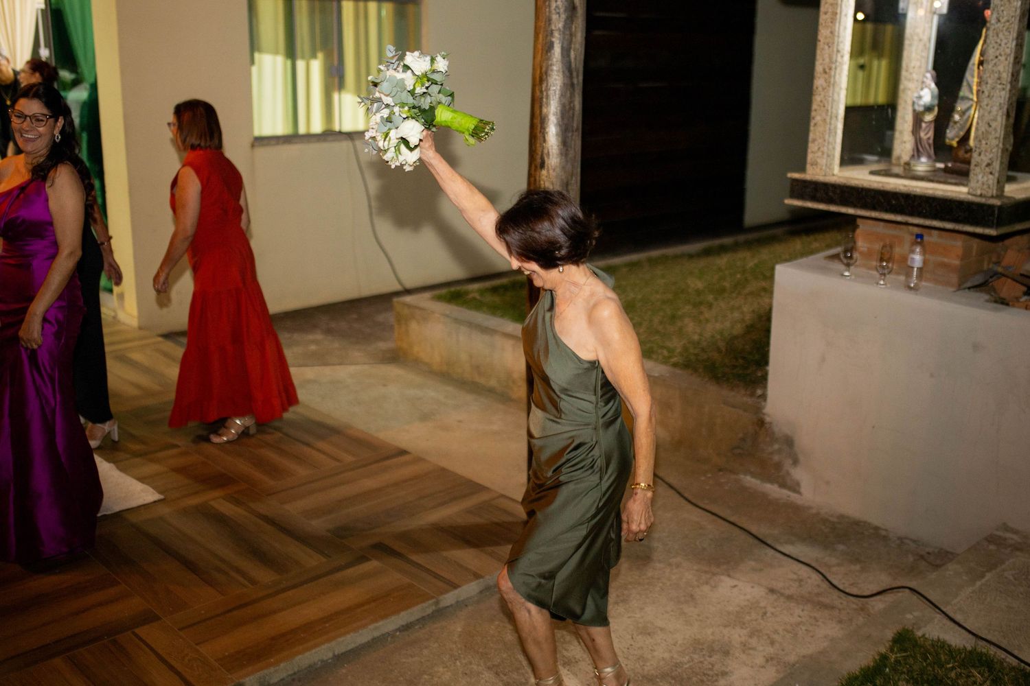 Woman in olive dress tossing wedding bouquet outdoors at night with guests in formal attire nearby.