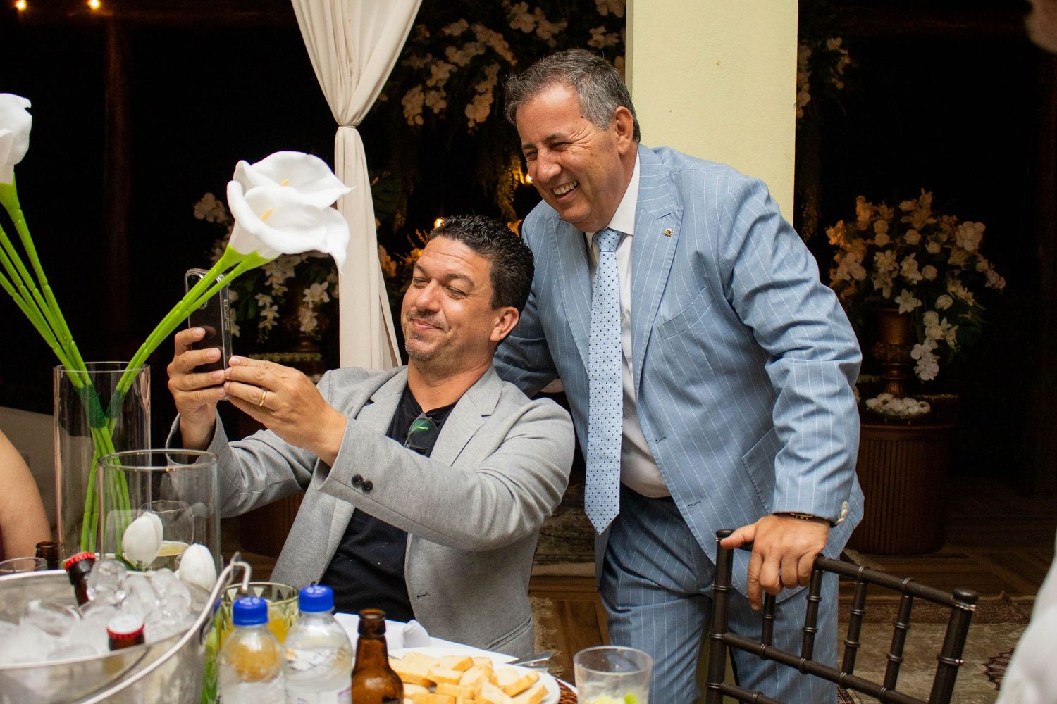 Two men in suits take a selfie at an outdoor evening event with drinks and food on the table nearby.