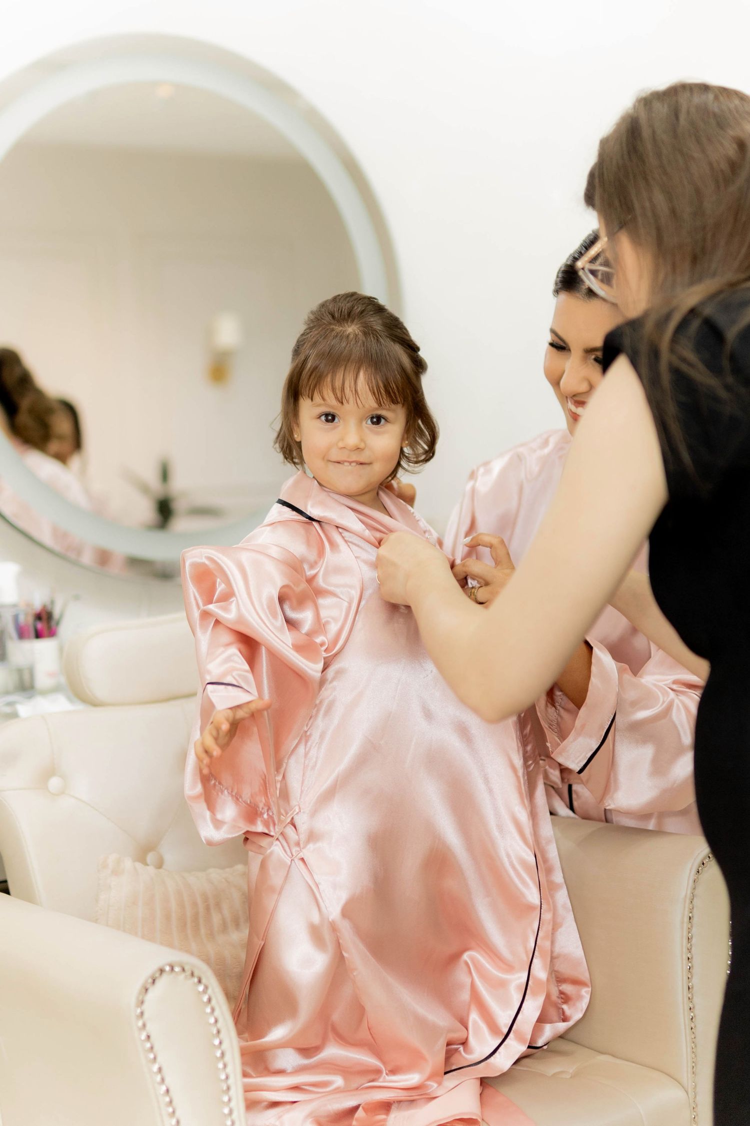 Young girl in pink satin robe being dressed by woman in bright salon with round mirror.