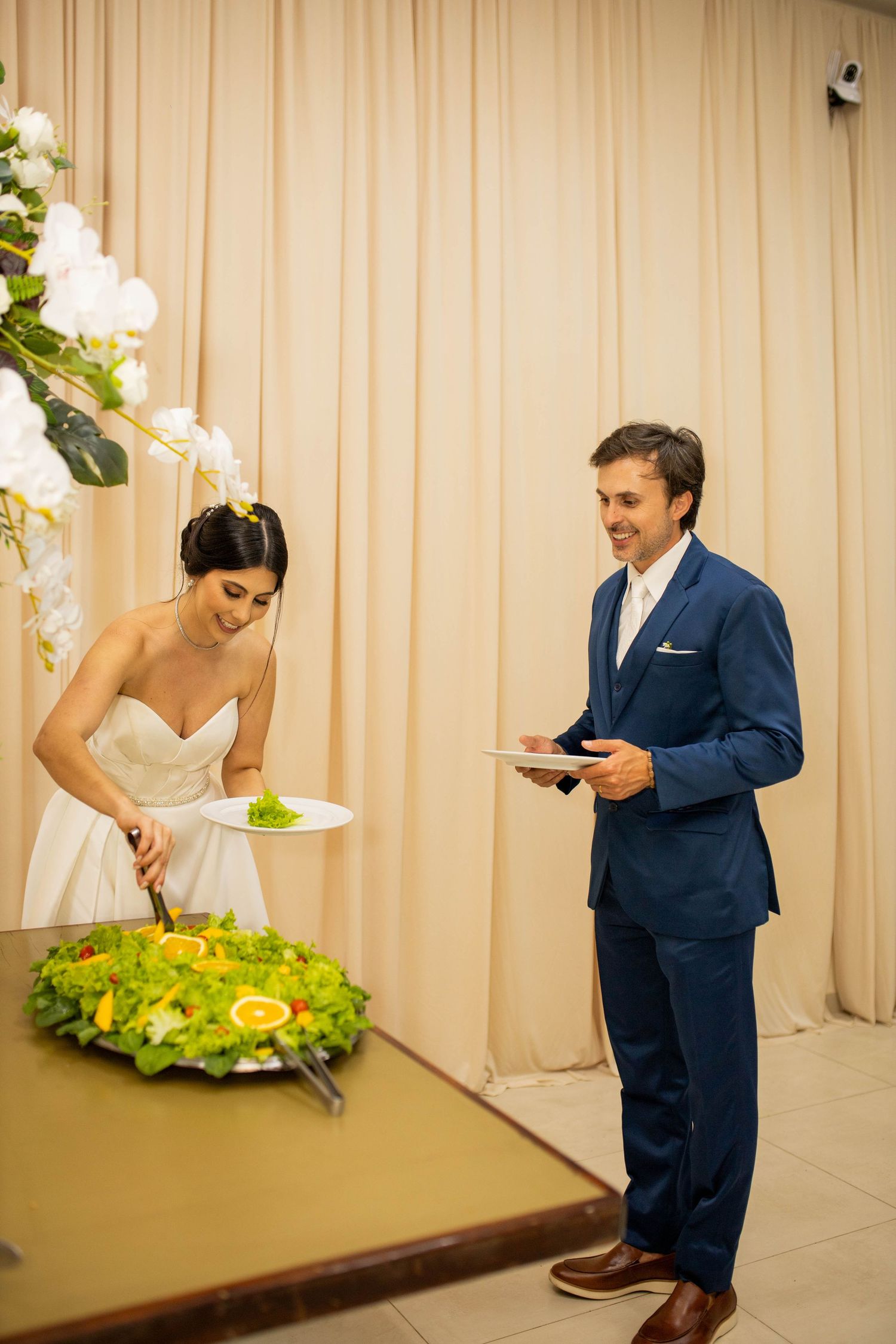 Bride in white gown cuts wedding cake while groom in navy suit holds plate at elegant reception venue.