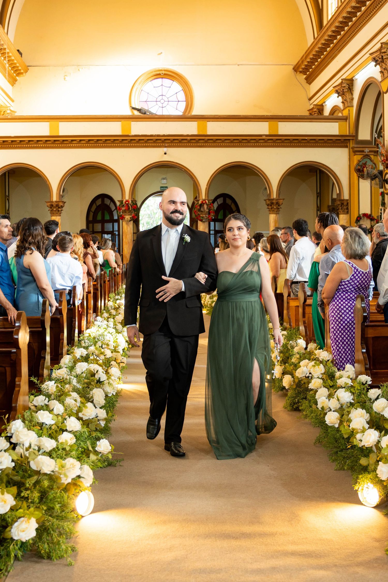 Groomsman in black tuxedo walks bridesmaid in dark green gown down decorated church aisle.