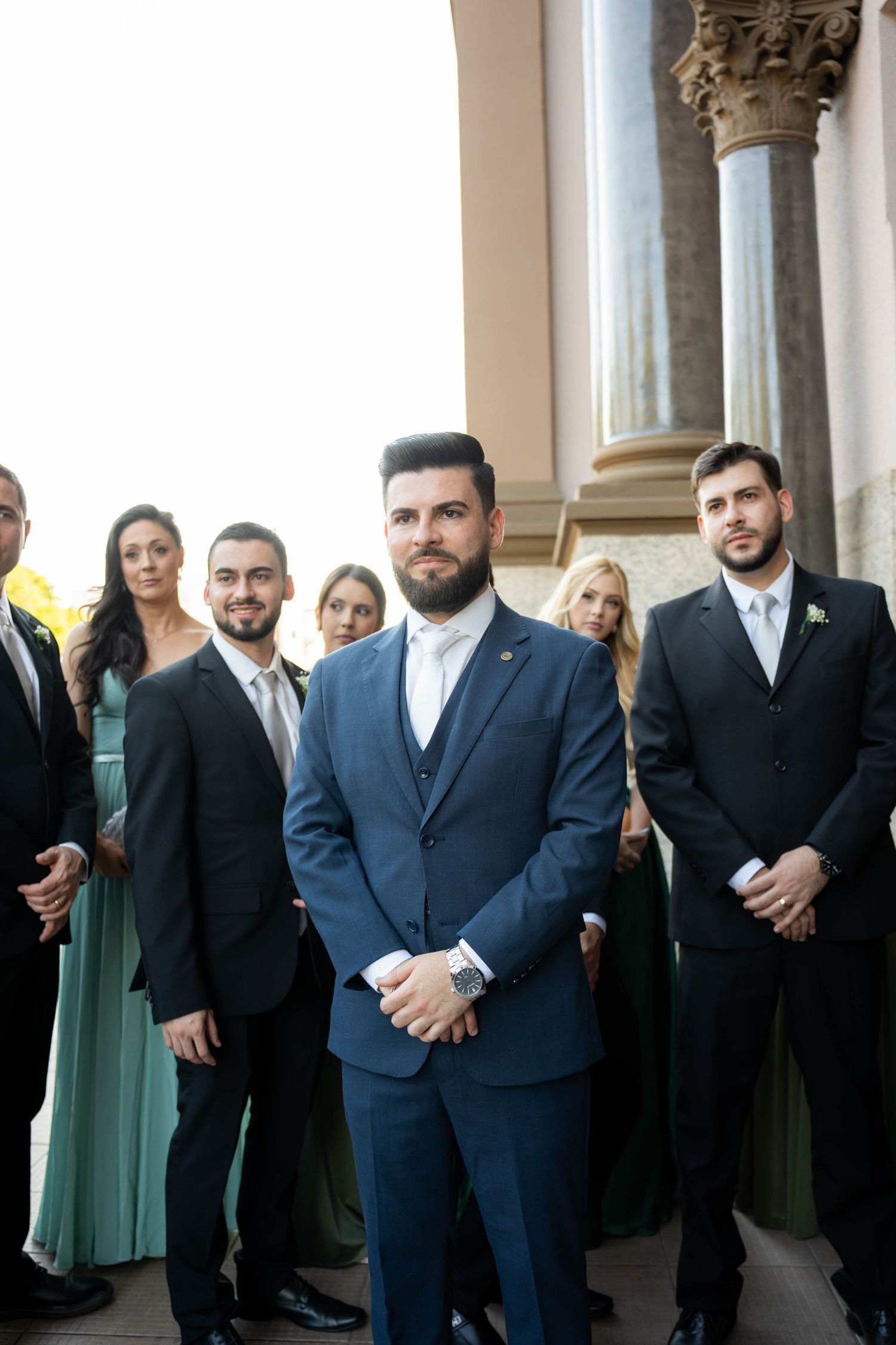 Groom in blue suit stands with wedding party in formal attire outside a classic building.