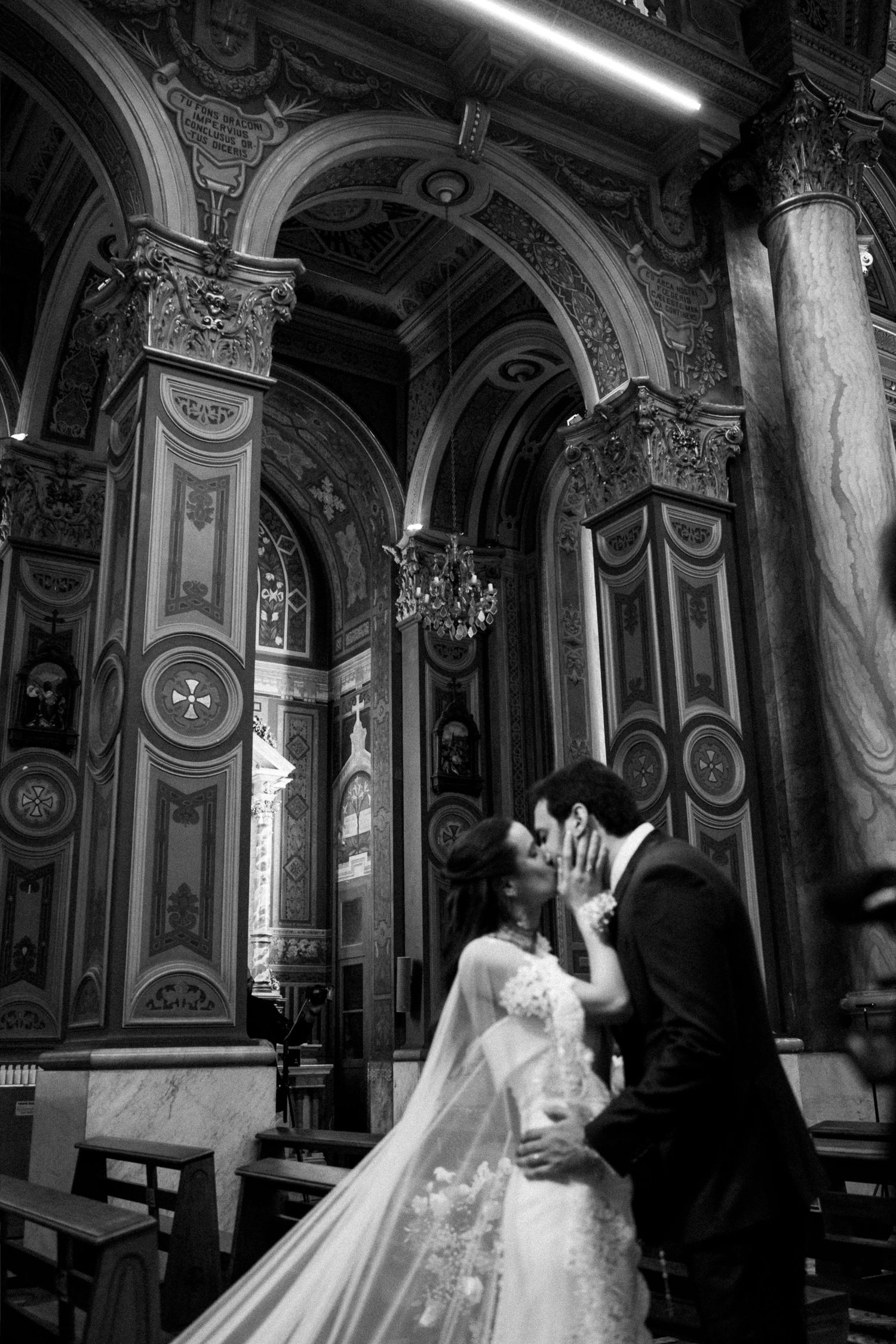 Black and white photo of a bride and groom kissing inside an ornate church with arched ceilings and chandelier.