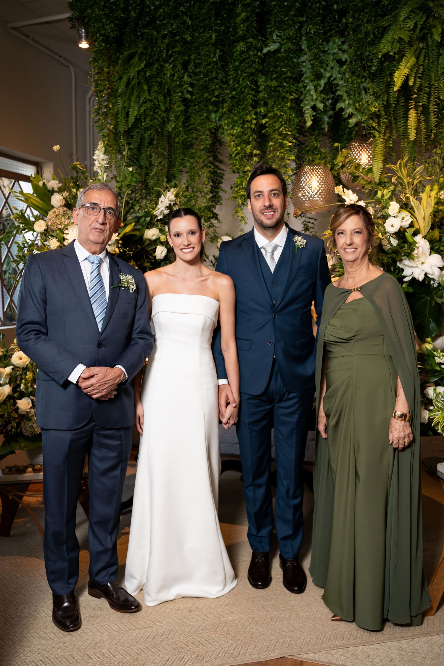 Wedding party of four posing in front of lush greenery backdrop with white floral arrangements.