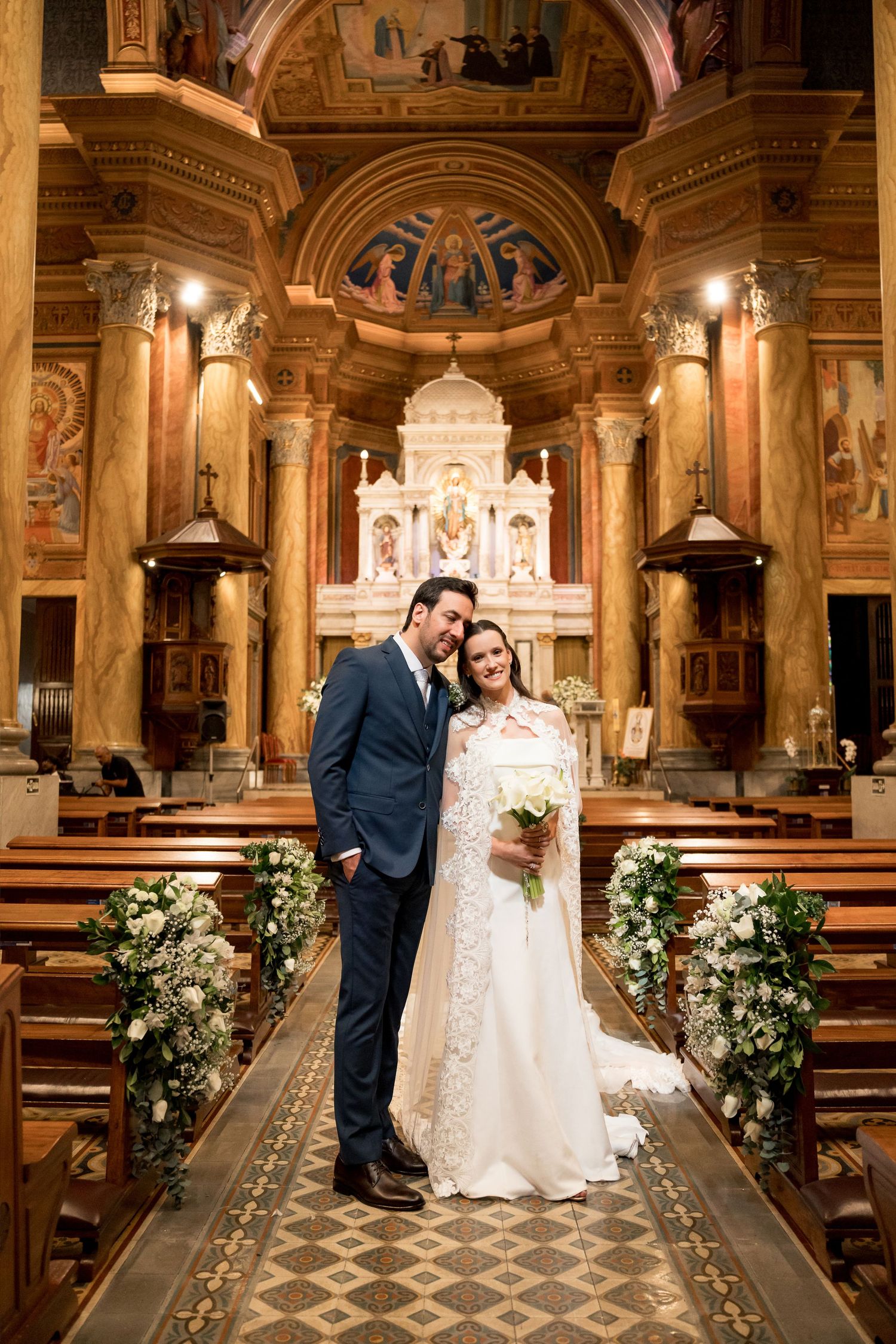 Bride and groom pose together in an ornate church aisle after their wedding ceremony, surrounded by floral arrangements.