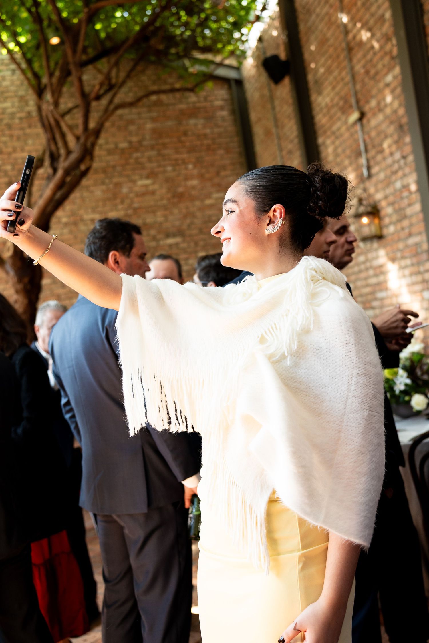 Young woman in white fringed shawl takes a selfie at an outdoor wedding reception near brick wall.