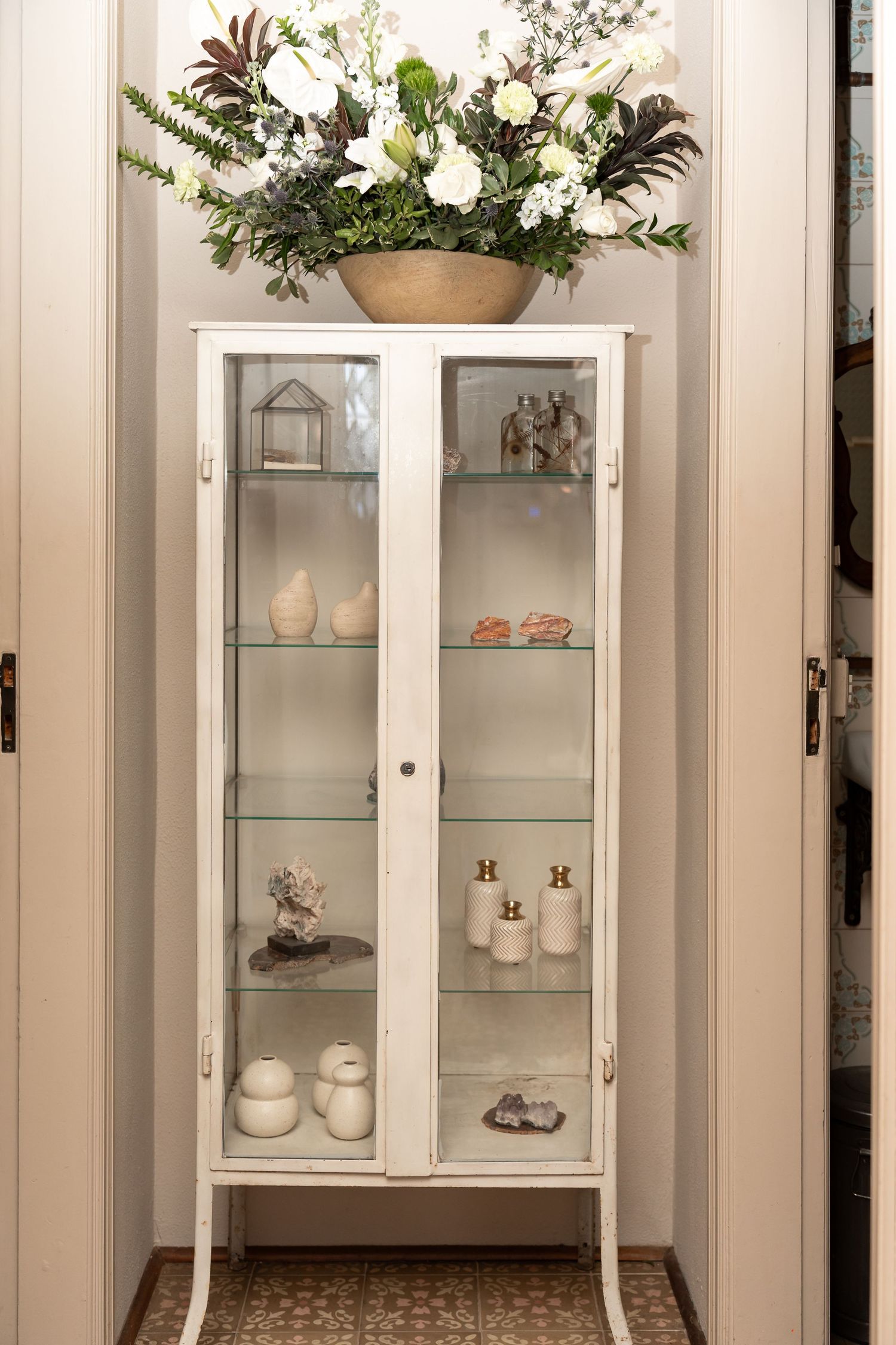 White glass display cabinet with decorative ceramics on shelves, topped with a floral arrangement in a wooden bowl.