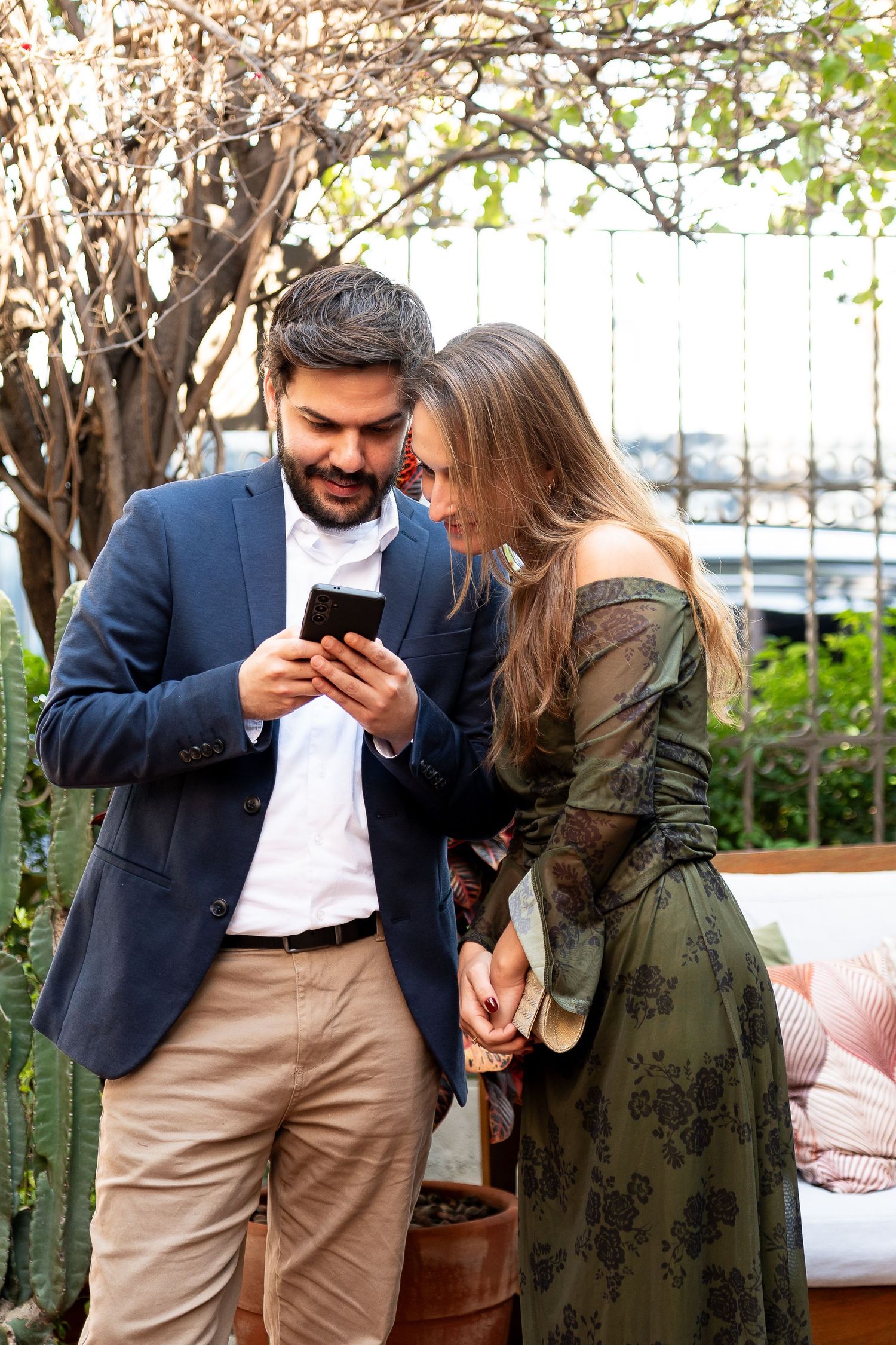 A smiling couple in elegant attire sharing a moment looking at a smartphone outdoors near lush greenery.