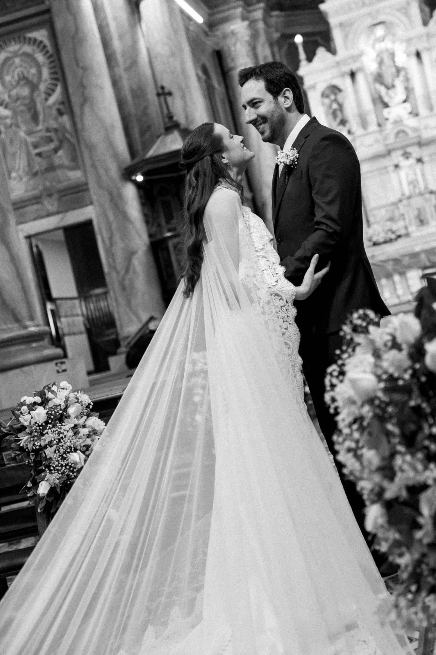 Black and white photo of a bride and groom sharing a joyful moment at the altar inside an elegant church.