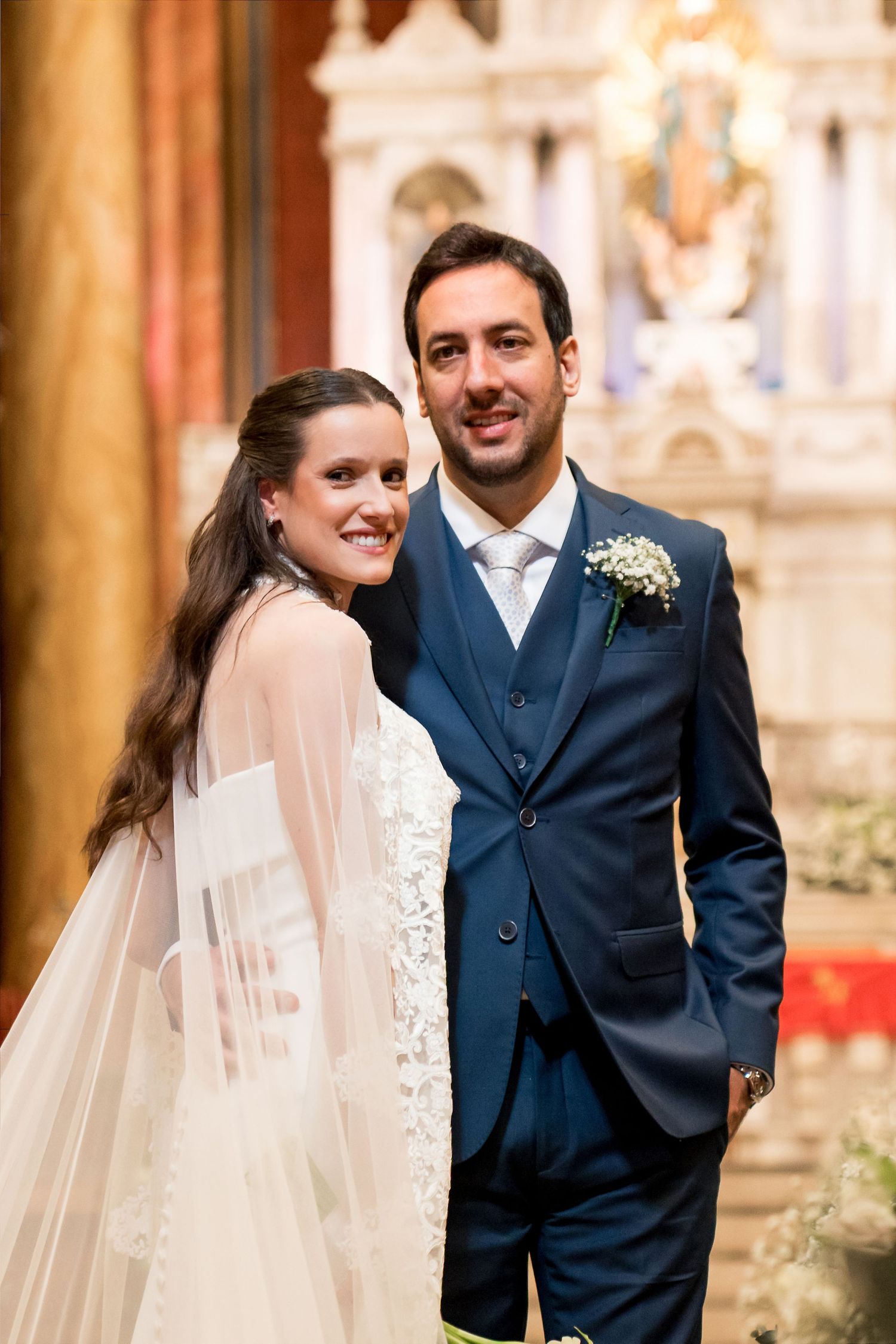 Bride in white lace gown and veil poses with groom in navy blue suit inside an ornate church.