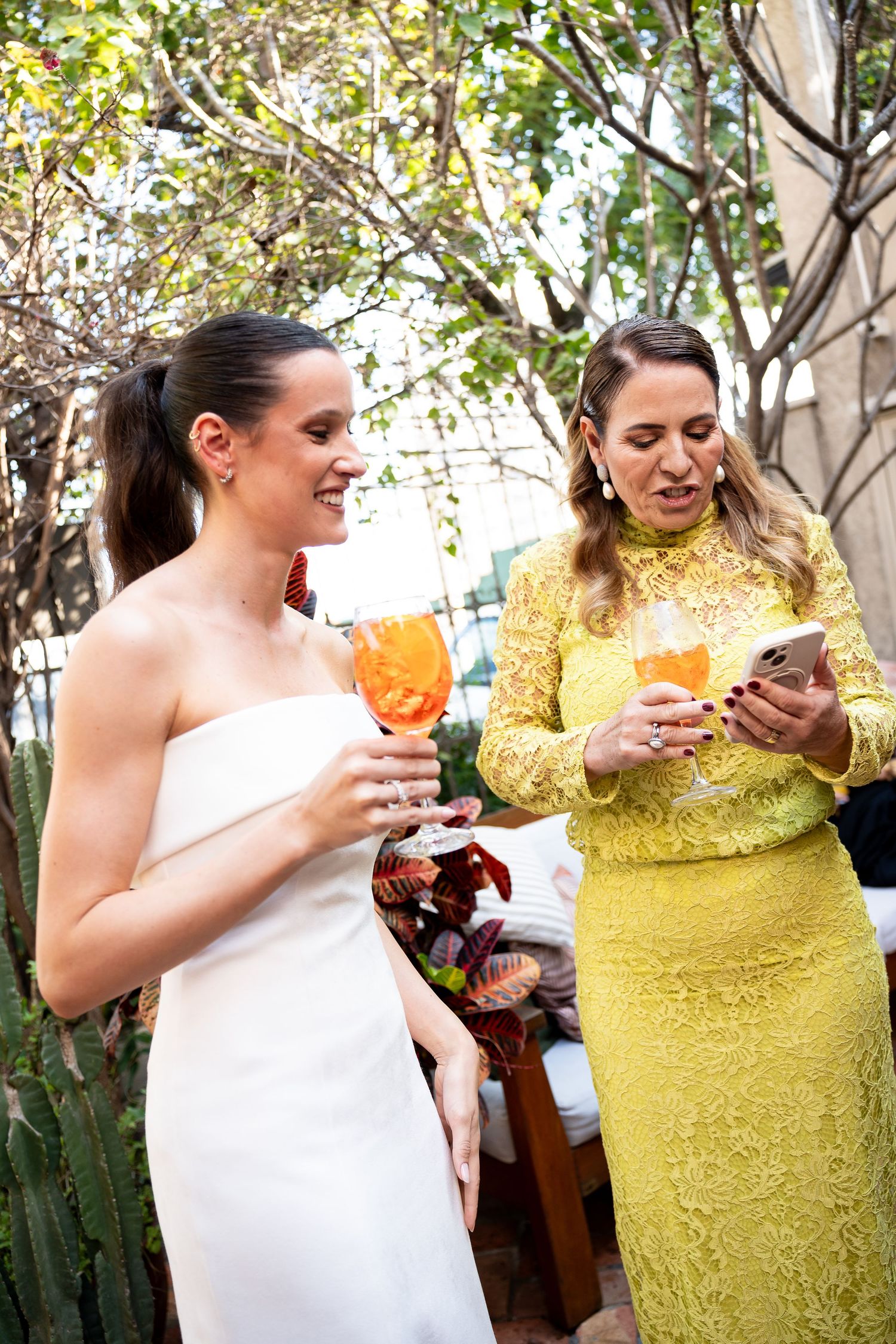 Two women holding orange cocktails outdoors, one in a white dress and one in a yellow lace dress checking her phone.