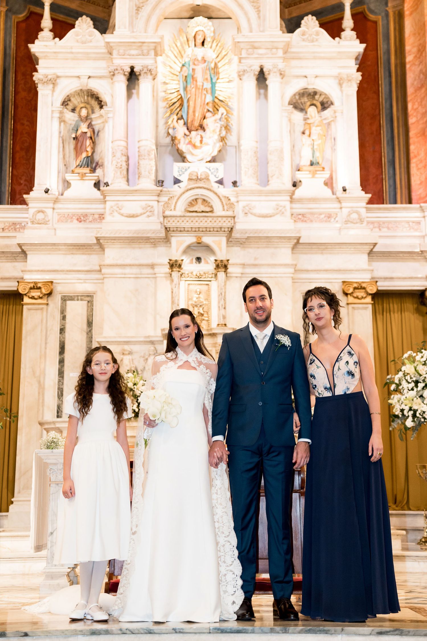 Wedding party poses at ornate church altar, bride in lace gown, groom in navy suit, bridesmaid and flower girl.