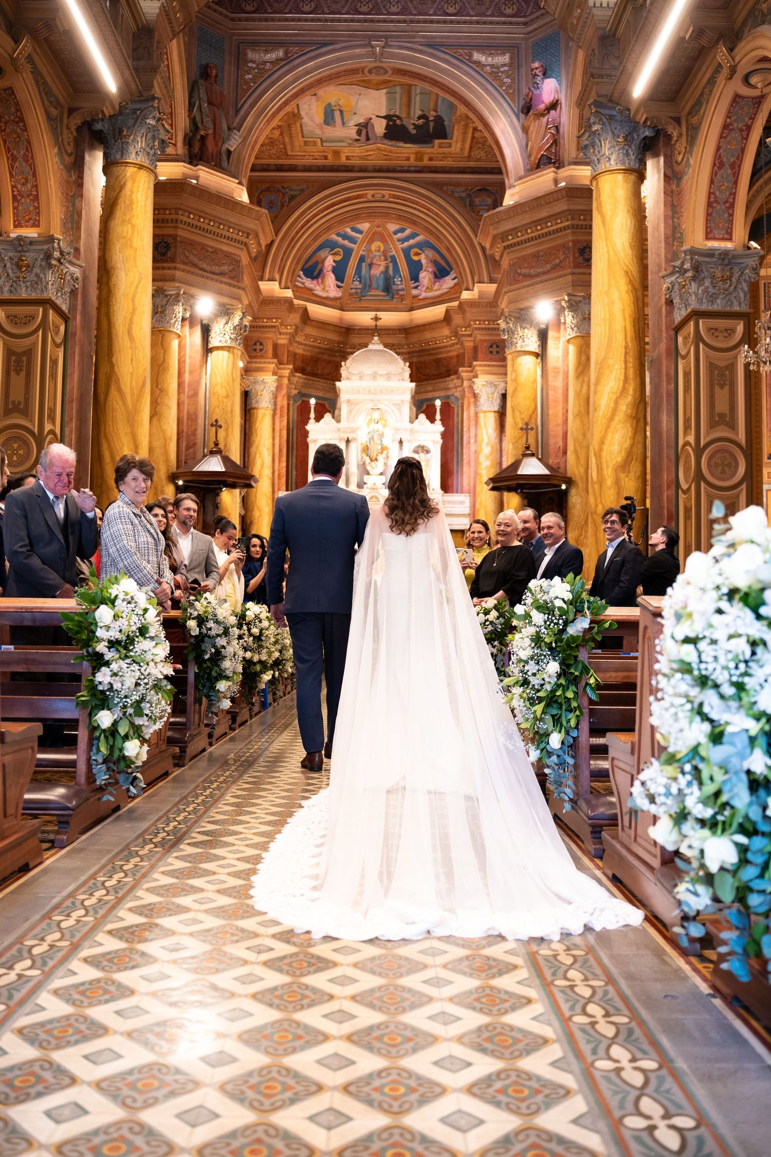 Bride and groom walking down the aisle of an ornate church decorated with white floral arrangements.