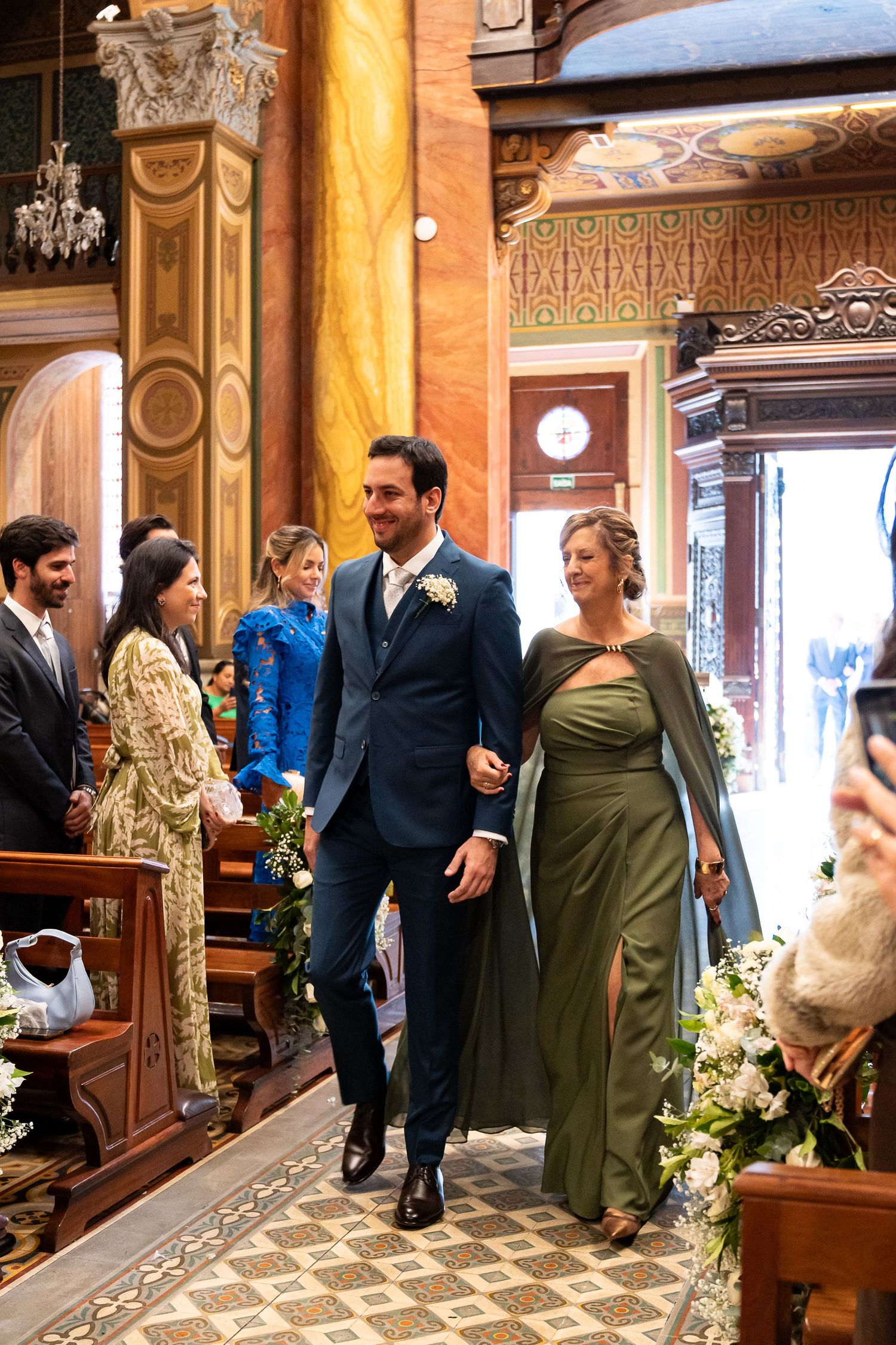 Groom in blue suit walks down church aisle with woman in green dress during elegant wedding ceremony.