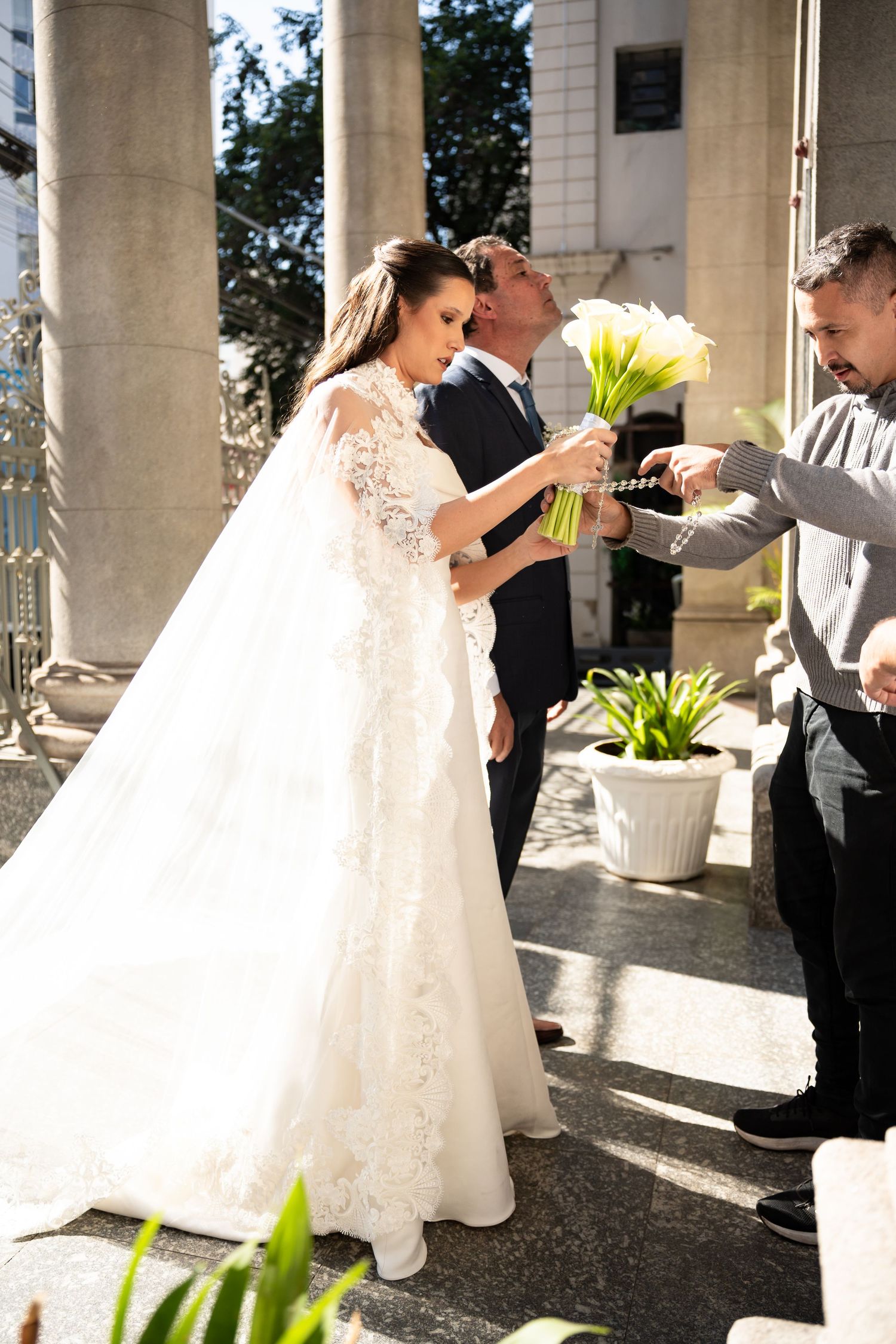 Bride in white lace gown receives bouquet of white calla lilies from guest, groom standing behind her outdoors.