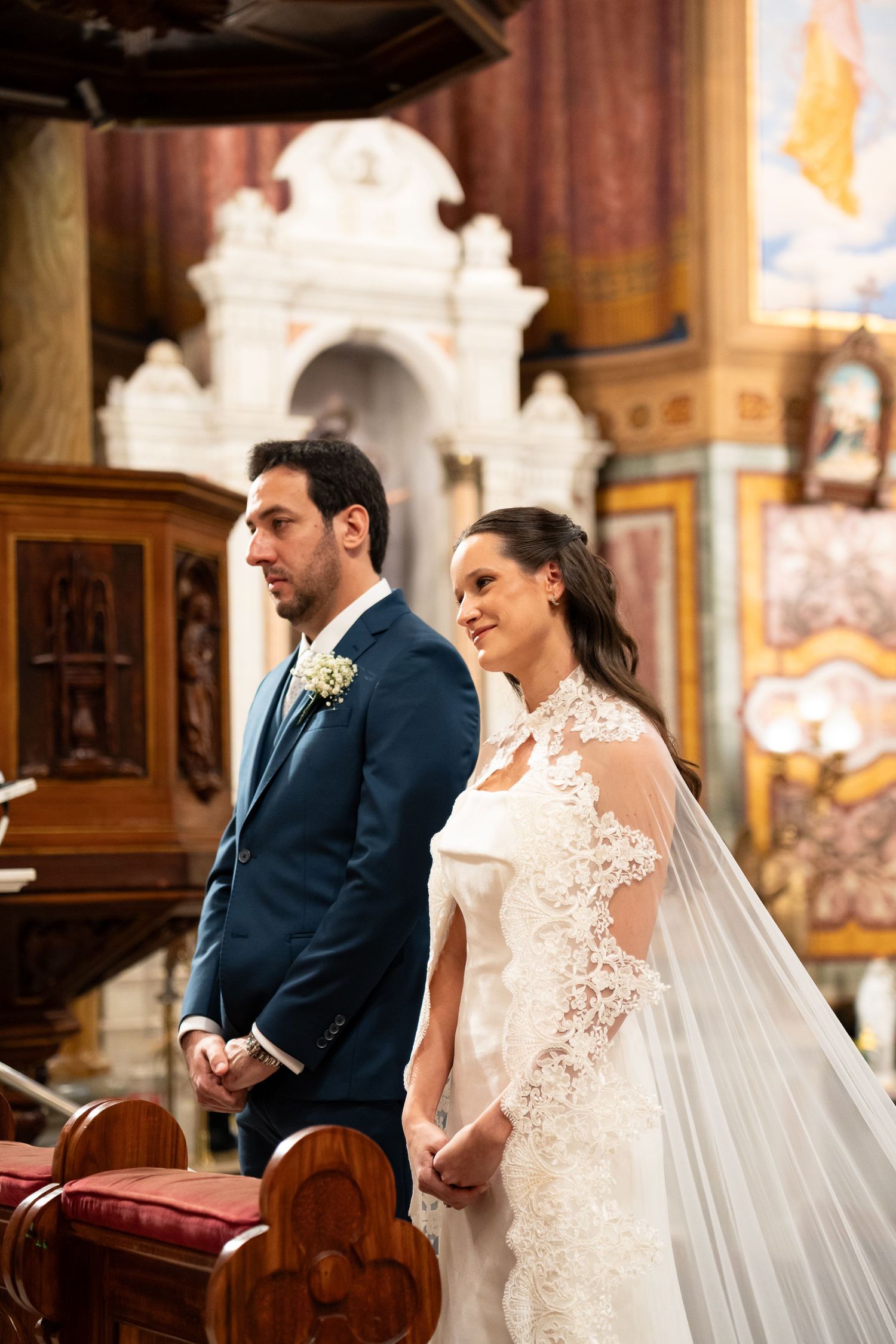 Bride in lace gown and groom in blue suit stand at church altar during wedding ceremony.