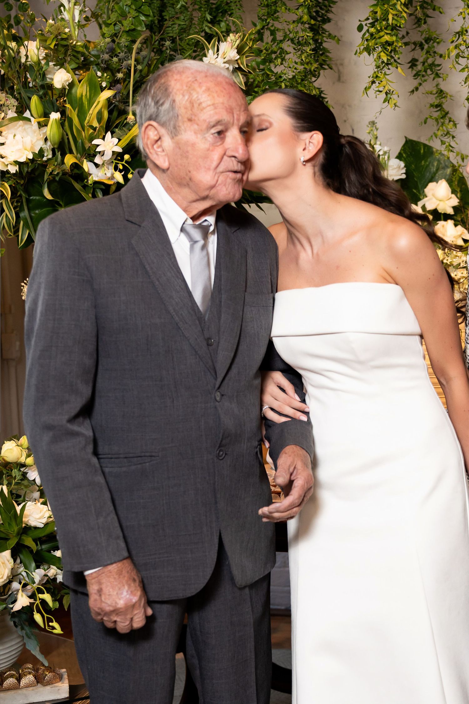 Bride in white strapless gown kisses elderly man in gray suit cheek at floral wedding ceremony backdrop.