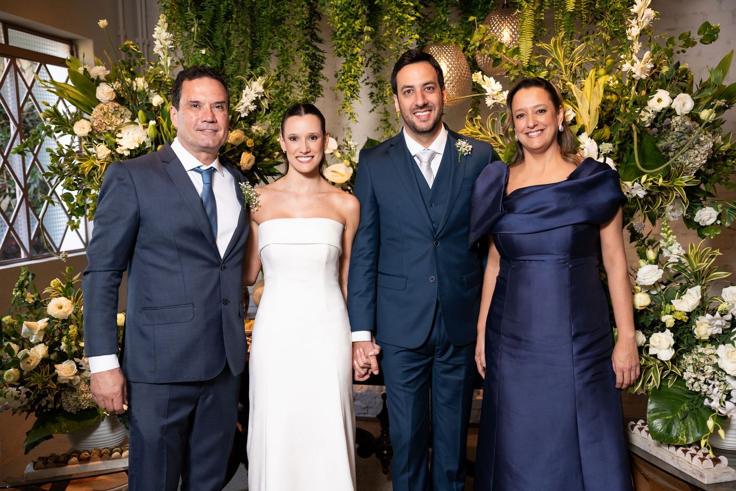 Wedding party of four posing with lush floral greenery backdrop, bride in white gown, groom in navy suit.