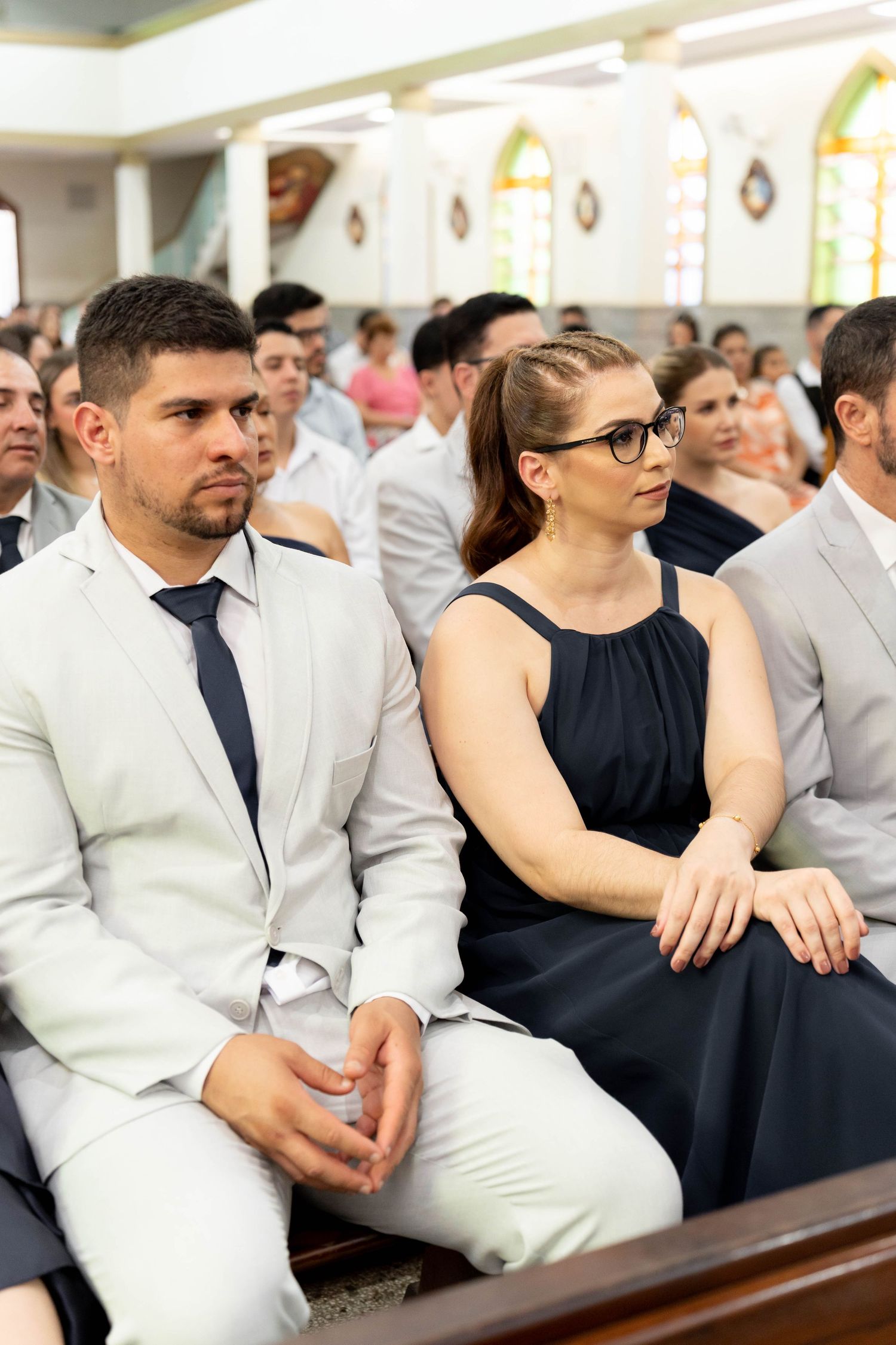 Elegantly dressed couple sitting in church pew during wedding ceremony, man in white suit, woman in black dress.