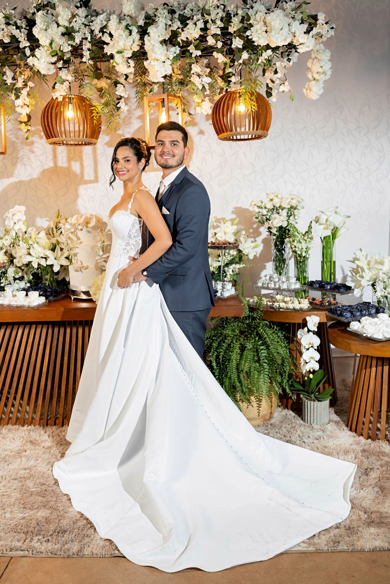Elegant bride and groom posing together at wedding reception with white floral decor and warm pendant lights.